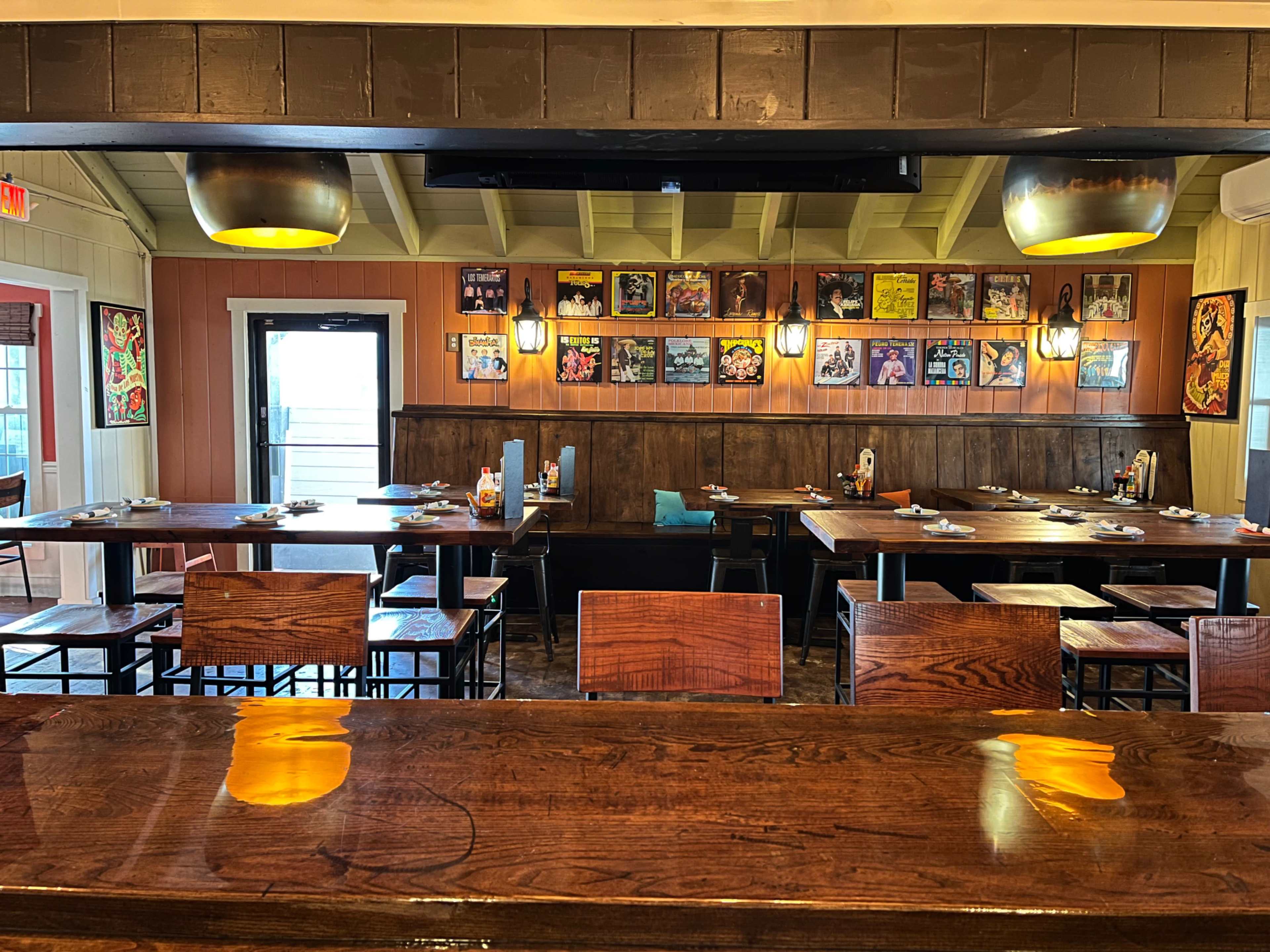 A restaurant interior with wooden tables and chairs, illuminated by pendant lights, and adorned with framed posters on the walls.