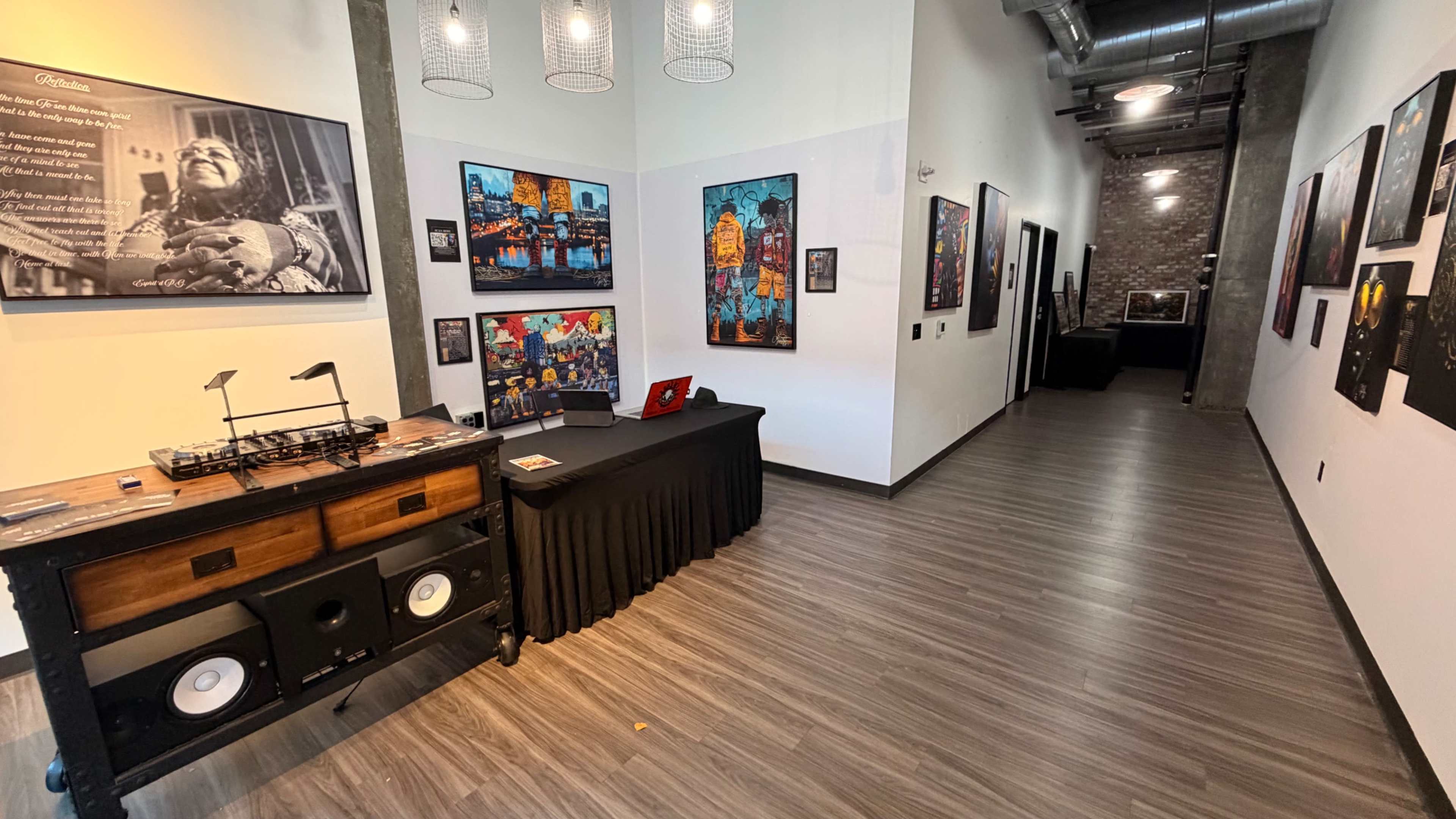 The image shows a hallway in an art gallery featuring framed artworks on the walls and a table with a DJ setup near the entrance.