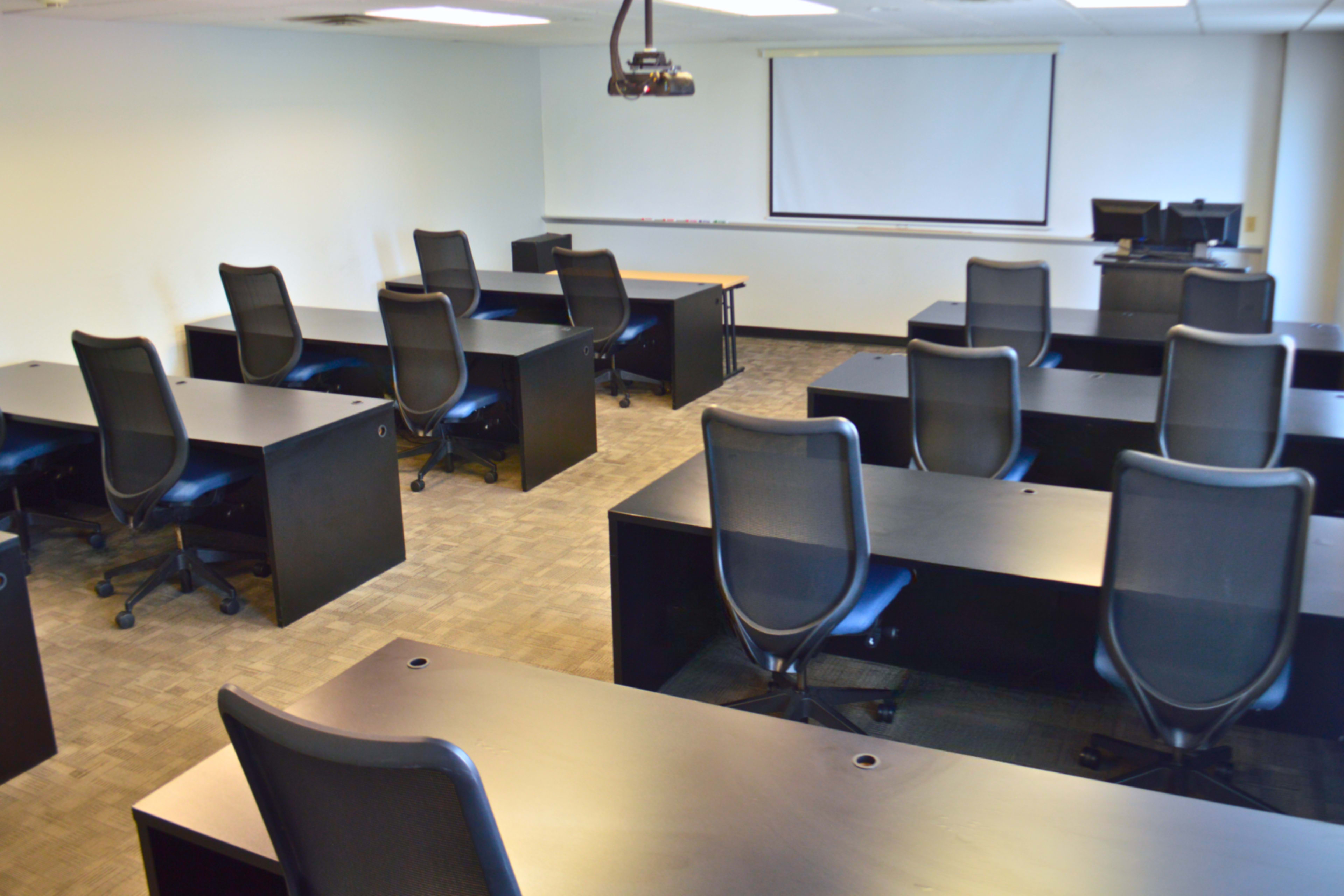 A classroom is set up with several rows of black desks and chairs, facing a projector screen and a computer station in the corner.