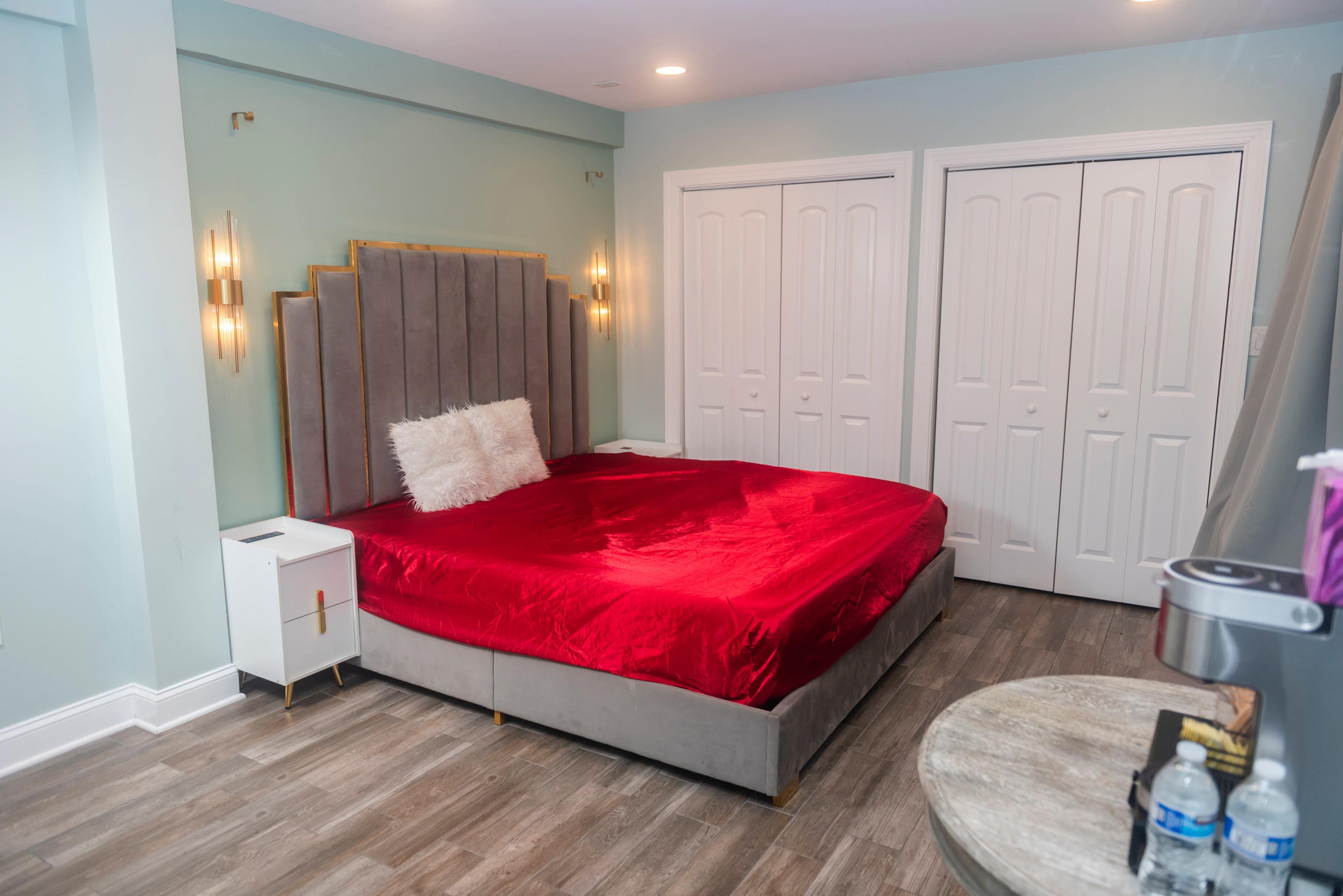 The image shows a bedroom featuring a large bed with a red duvet, a decorative headboard, and two white closet doors.