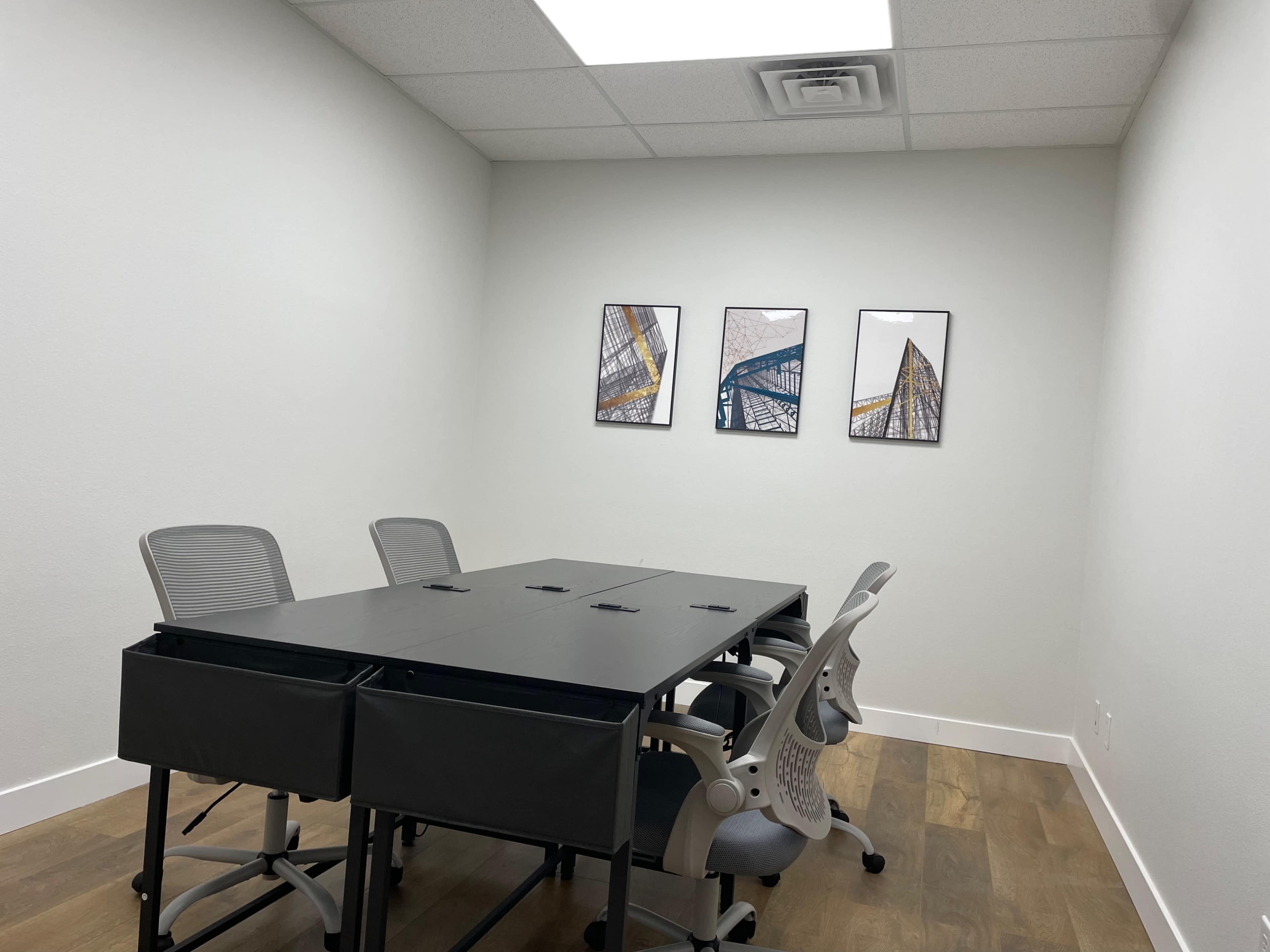 A small conference room with a rectangular table and four ergonomic chairs, featuring three framed artworks on the wall.