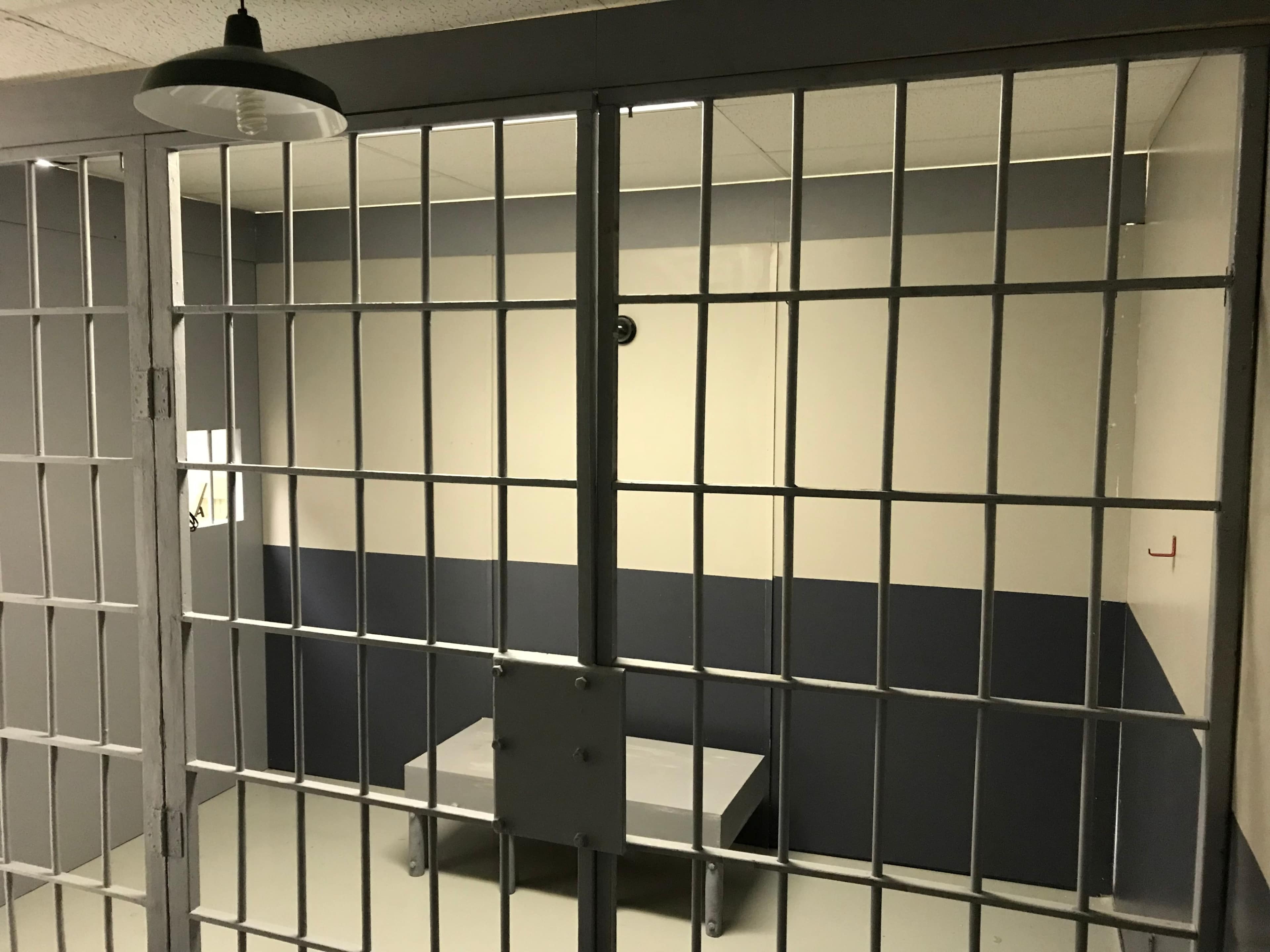 The image shows an empty jail cell with barred doors and a plain table against the wall.