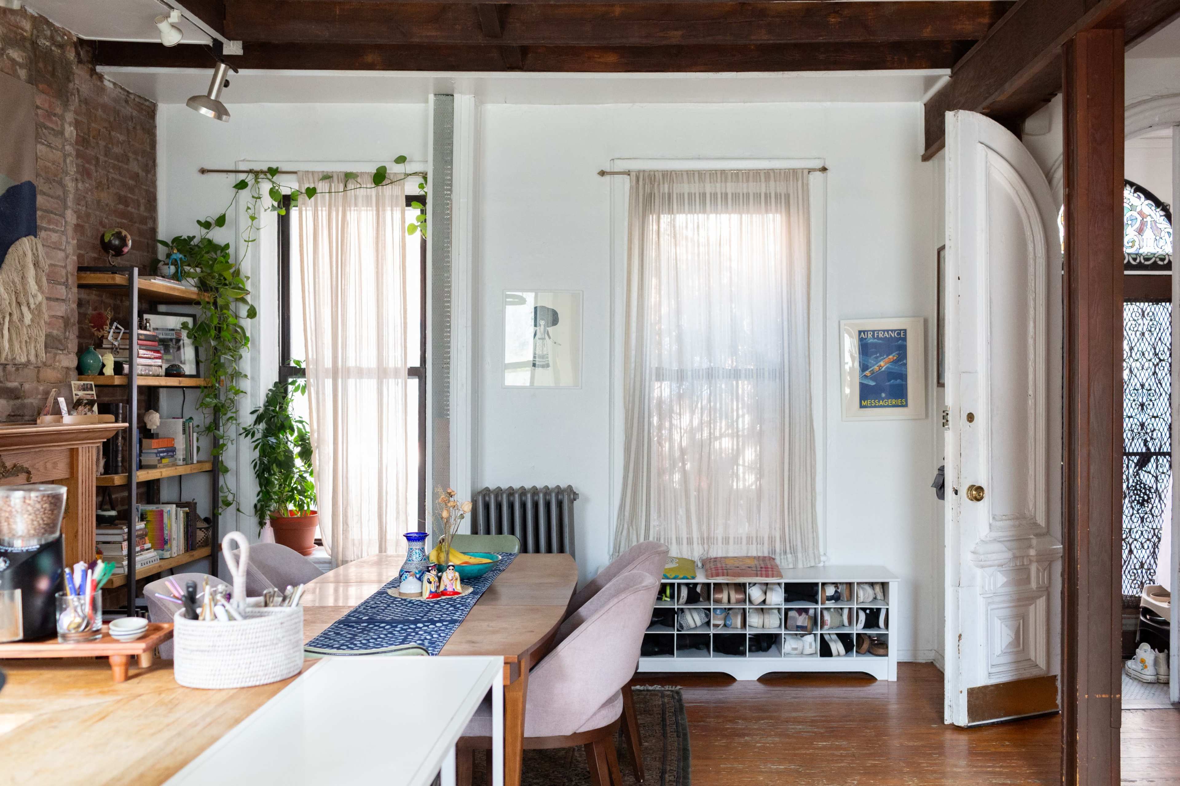 A bright room features a wooden dining table, light pink chairs, a plant near the window, and shelves filled with books and decor.