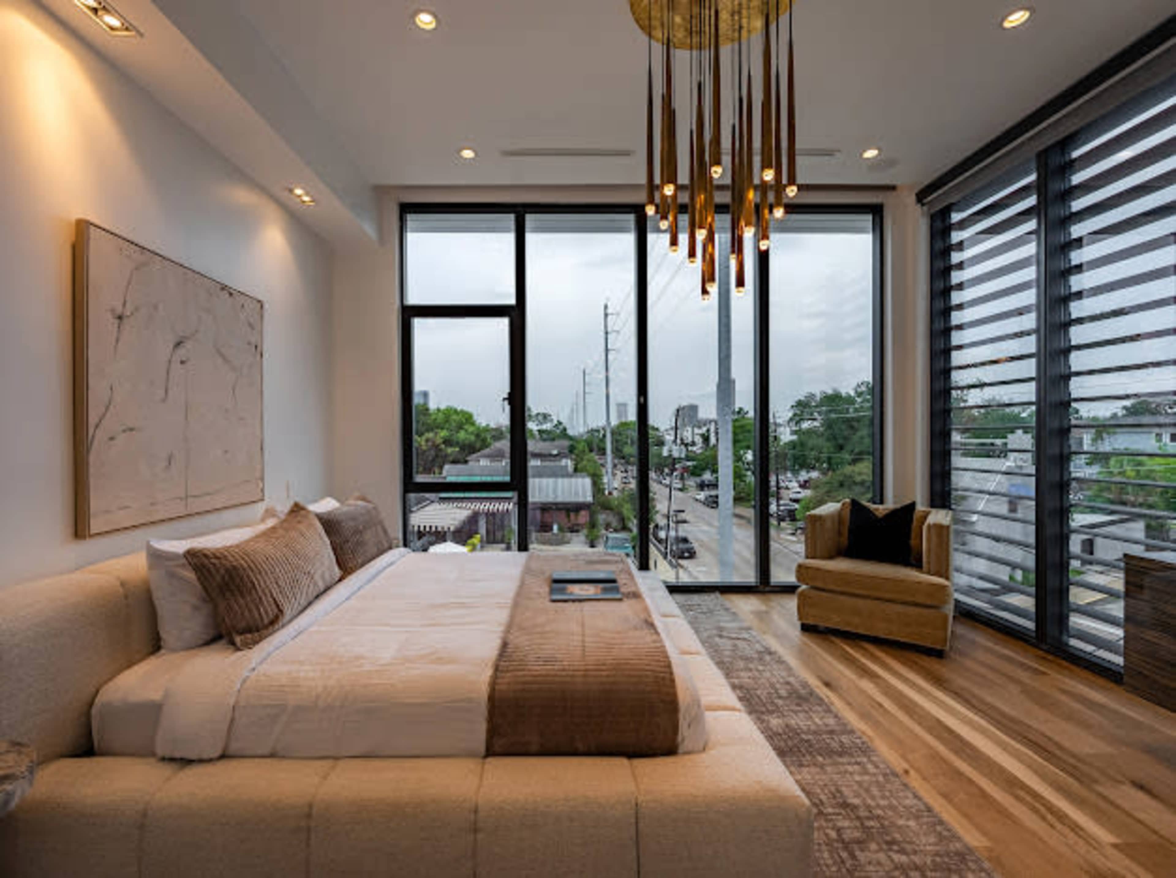 A modern bedroom features a large bed with a neutral color scheme, large windows overlooking a street, and stylish pendant lighting.