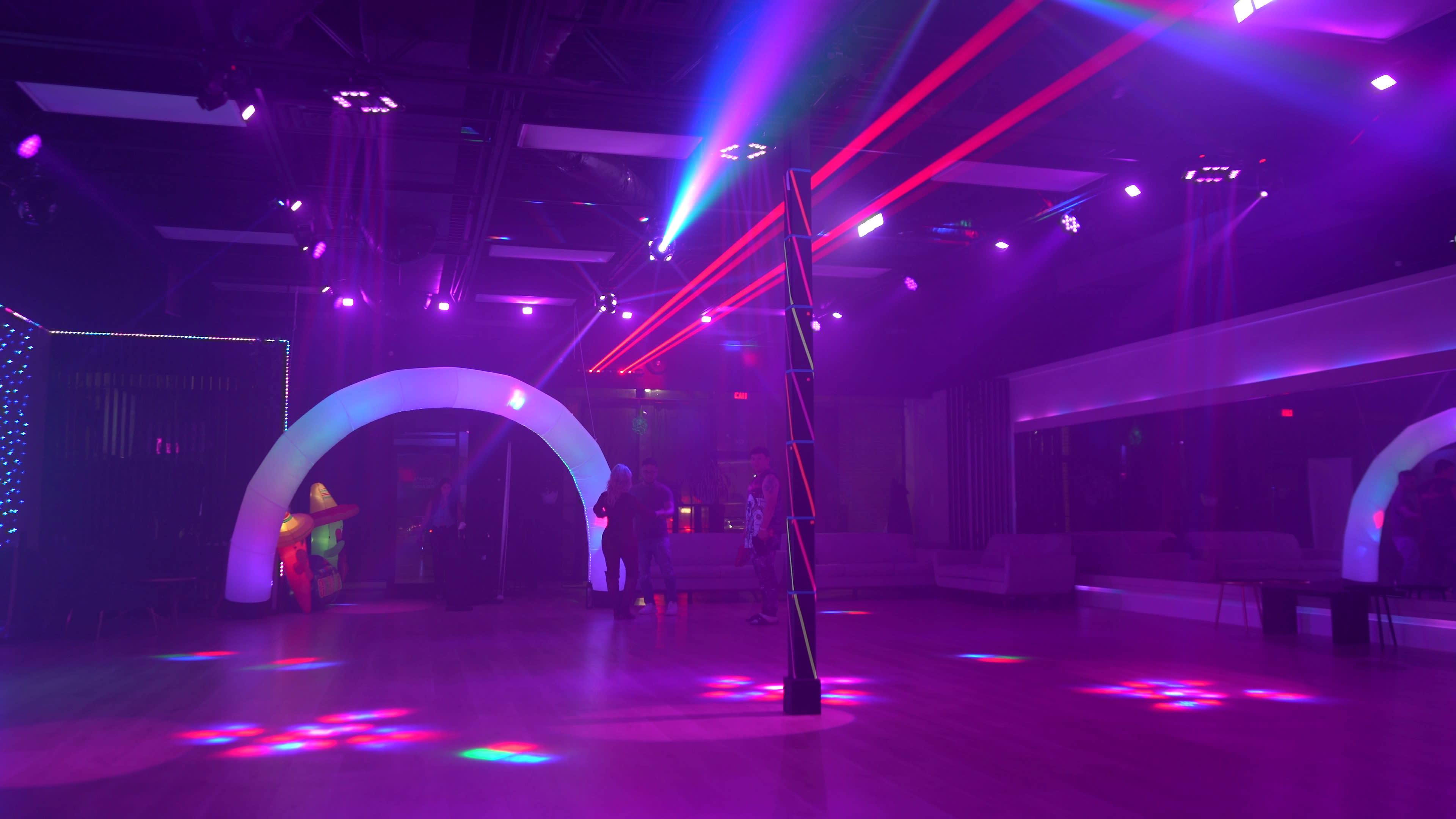 The image shows a dimly lit dance floor with colorful laser lights and an illuminated arch structure in a modern venue.