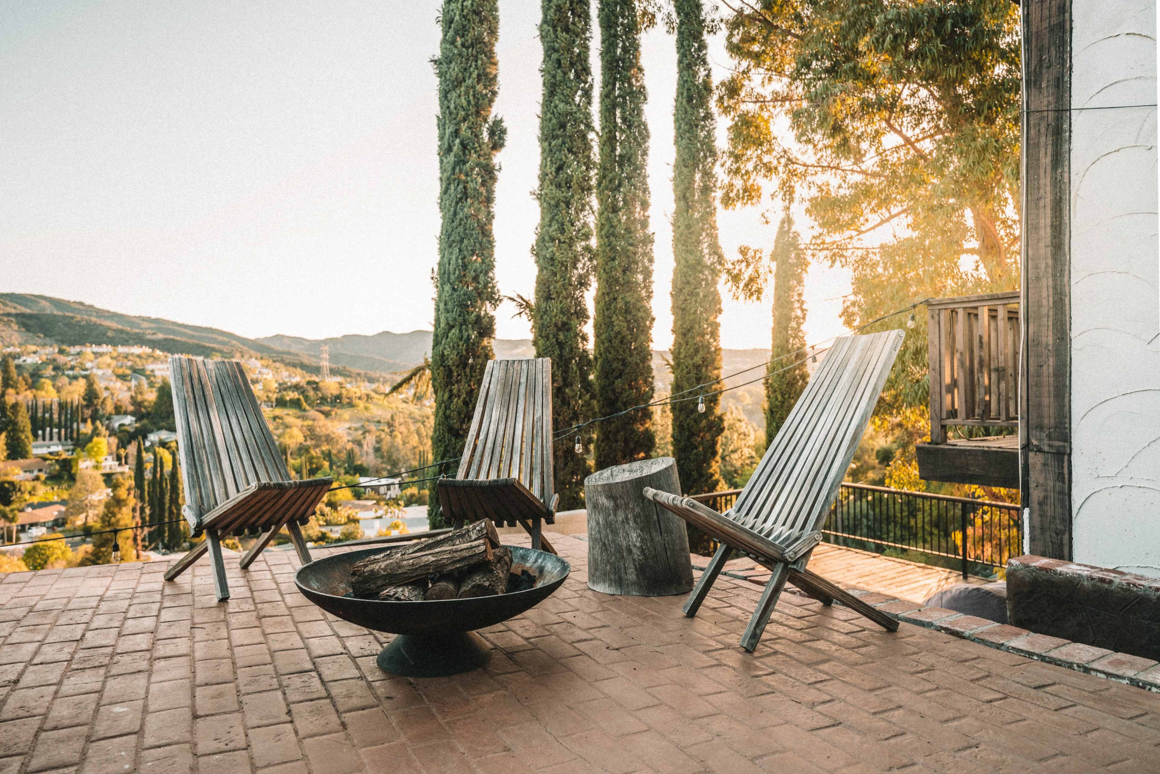 A seating area on a patio with two wooden chairs facing a fire pit, surrounded by tall trees and hills in the background.
