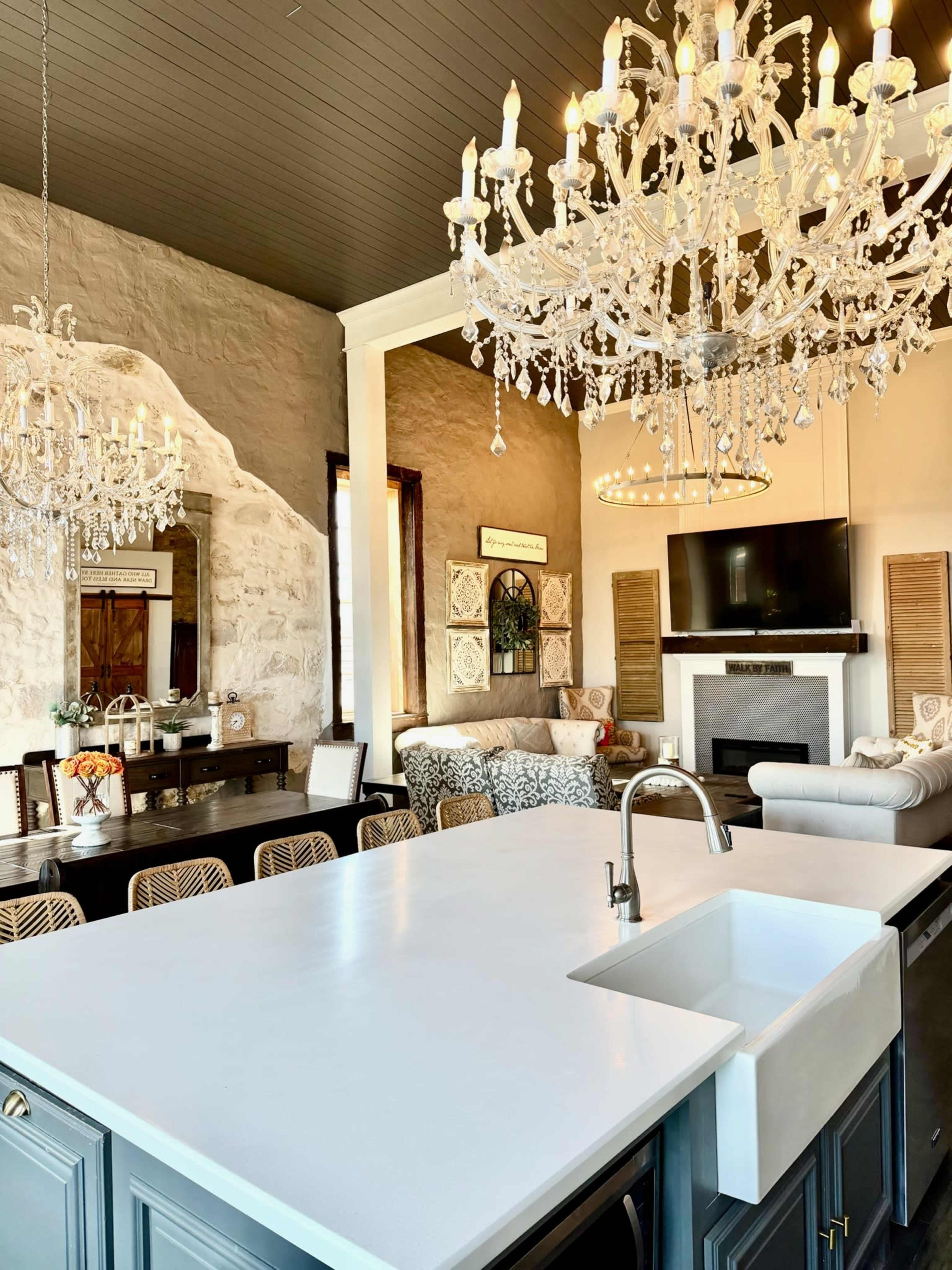 The image shows a modern living and dining area featuring a large kitchen island, multiple chandeliers, and a stylish seating arrangement.