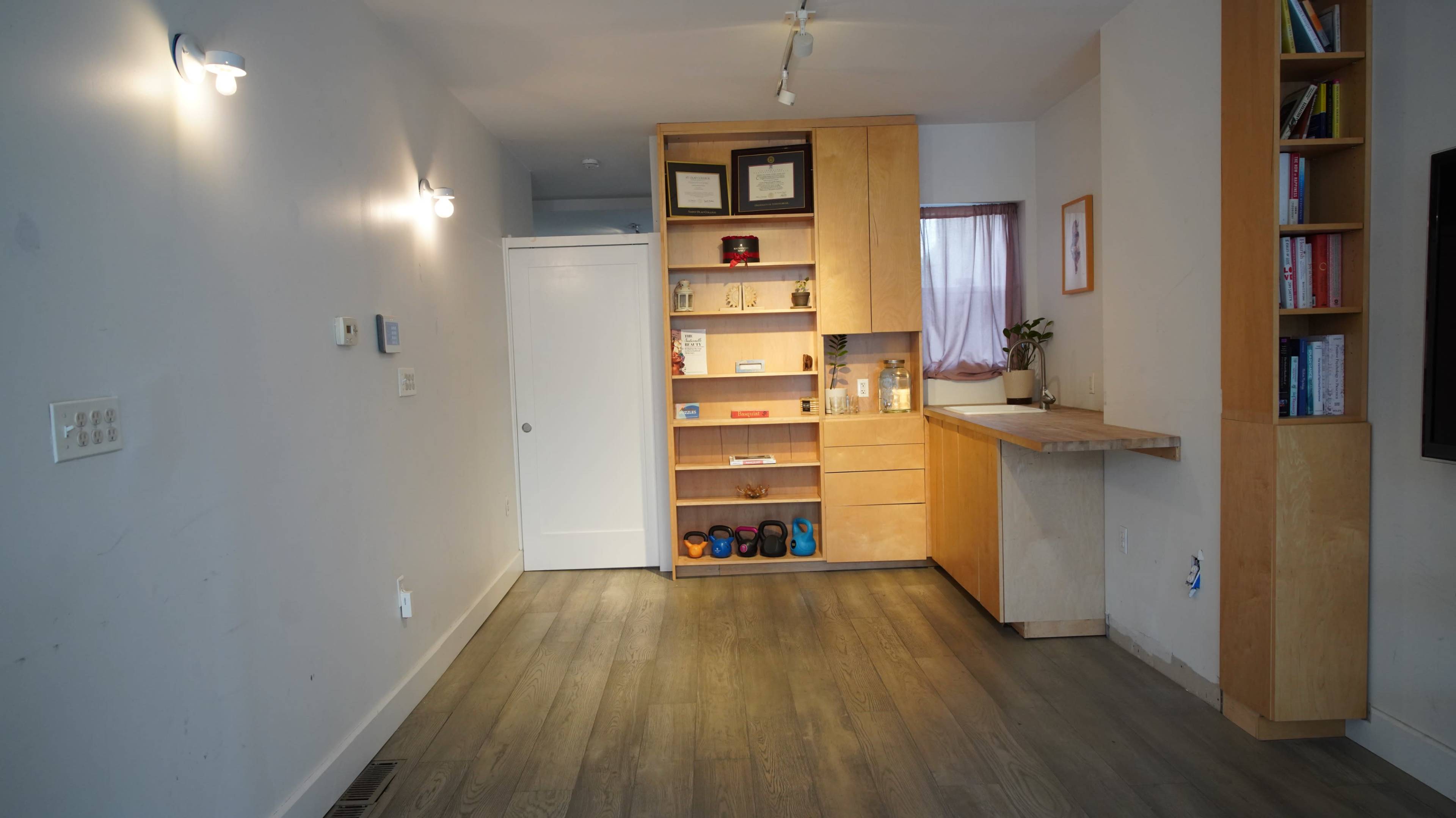 The image shows a minimalist interior space featuring a wooden shelving unit with various items and a small counter, along with light gray walls and wooden flooring.