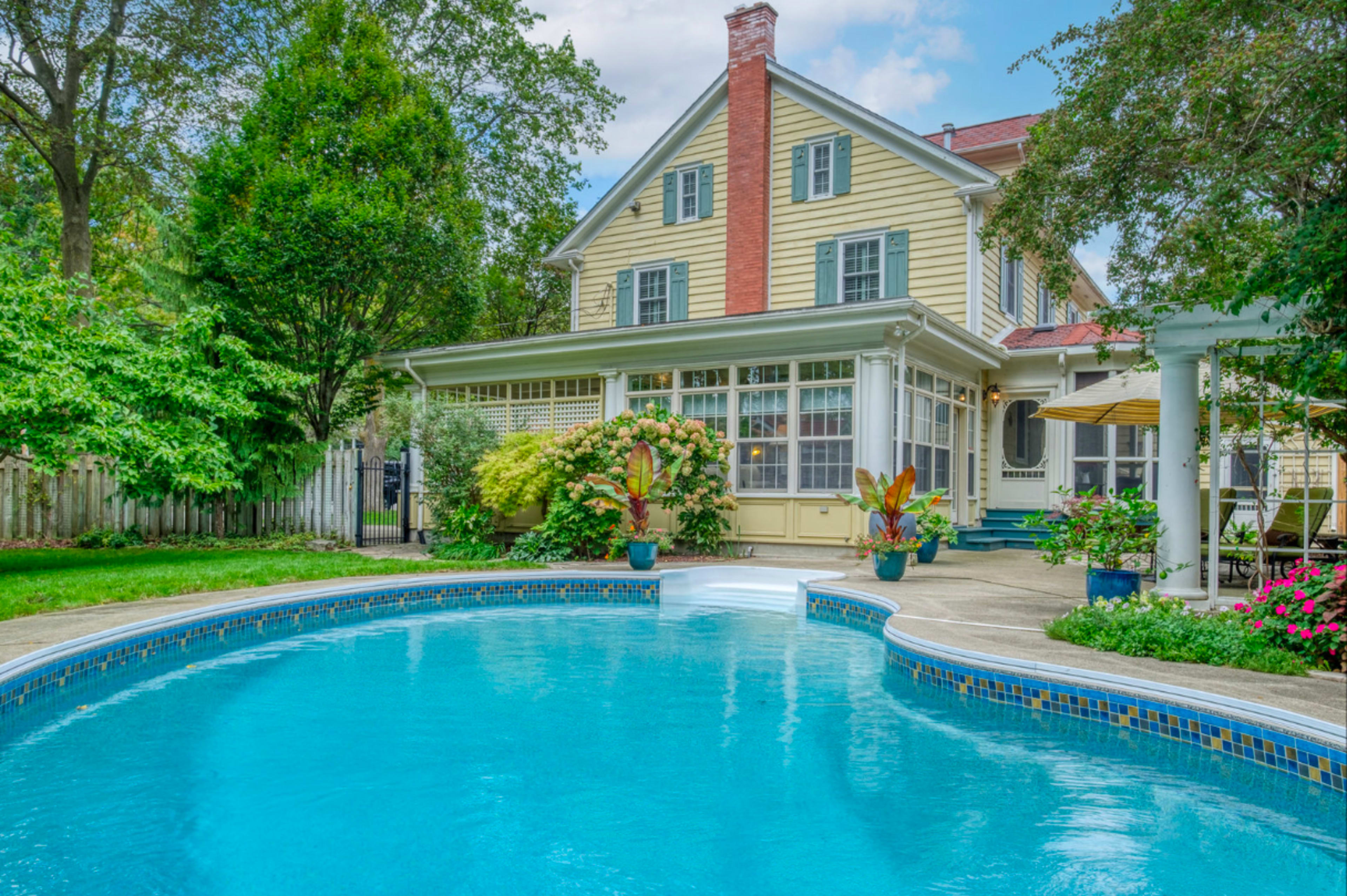 A backyard with a blue swimming pool in front of a two-story yellow house surrounded by lush greenery and colorful flower pots.