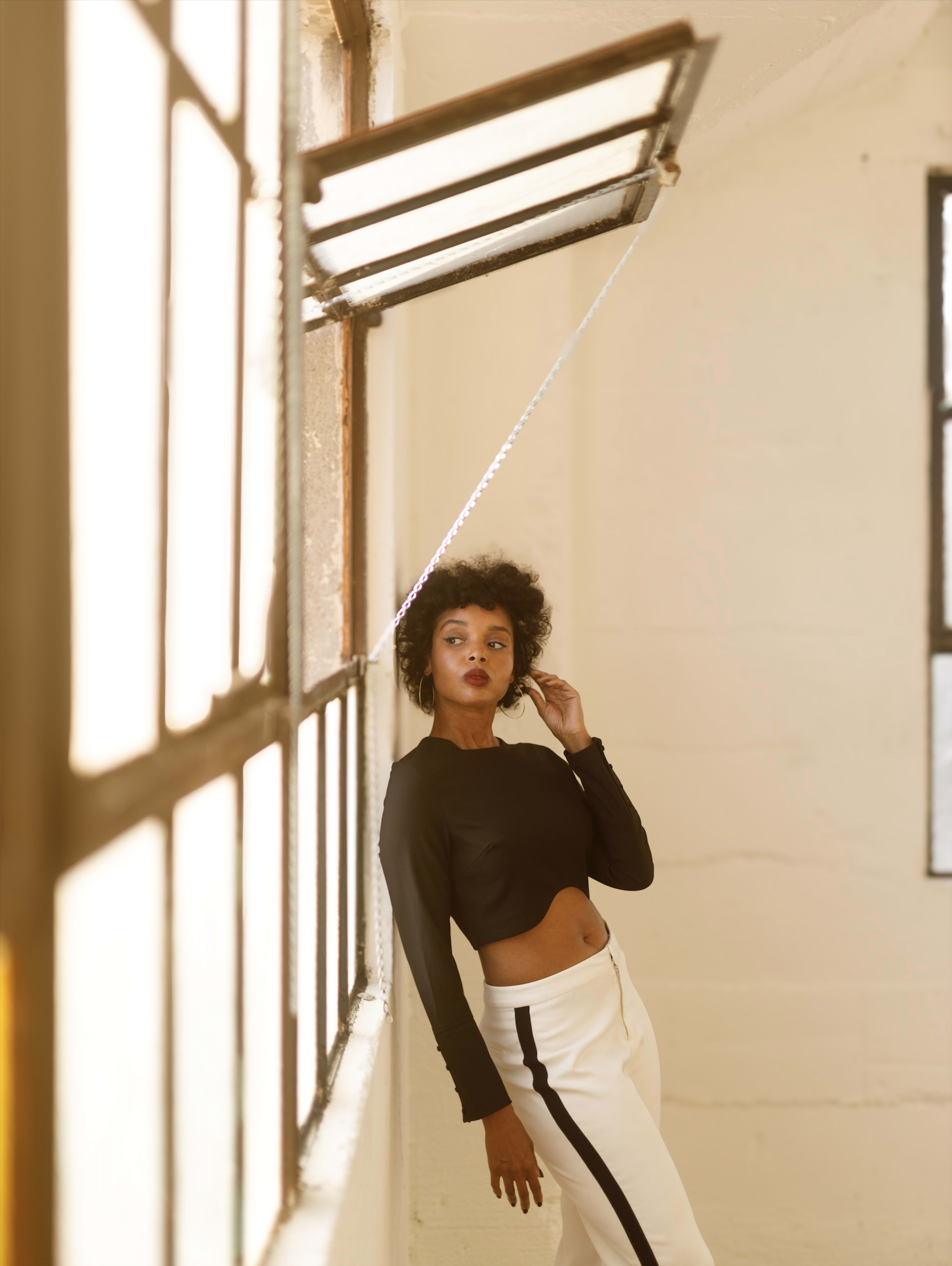 A woman with curly hair poses in a white room next to an open window, wearing a black crop top and white pants.