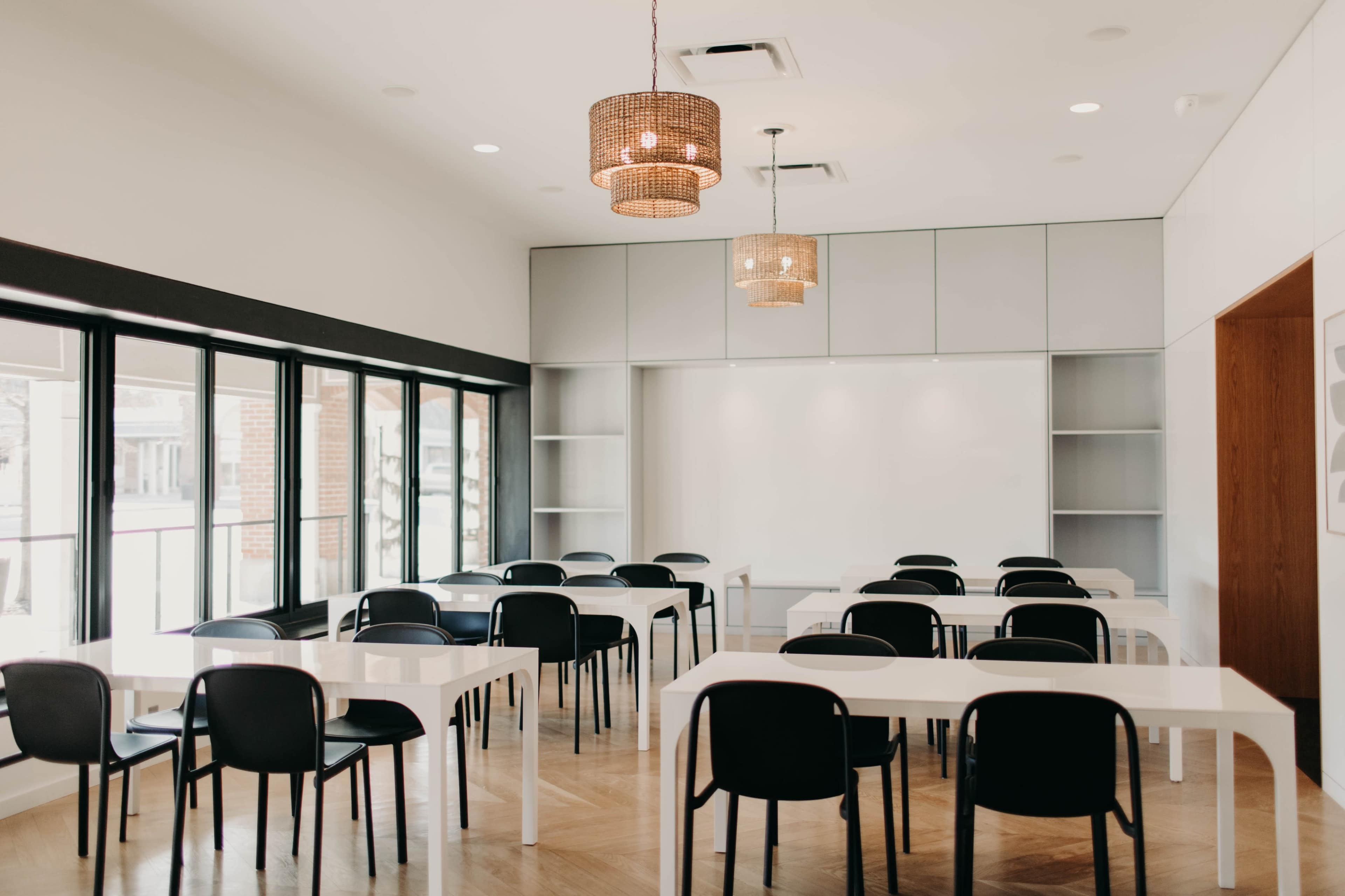 A bright conference room features multiple rectangular tables and black chairs, with large windows allowing natural light to illuminate the space.