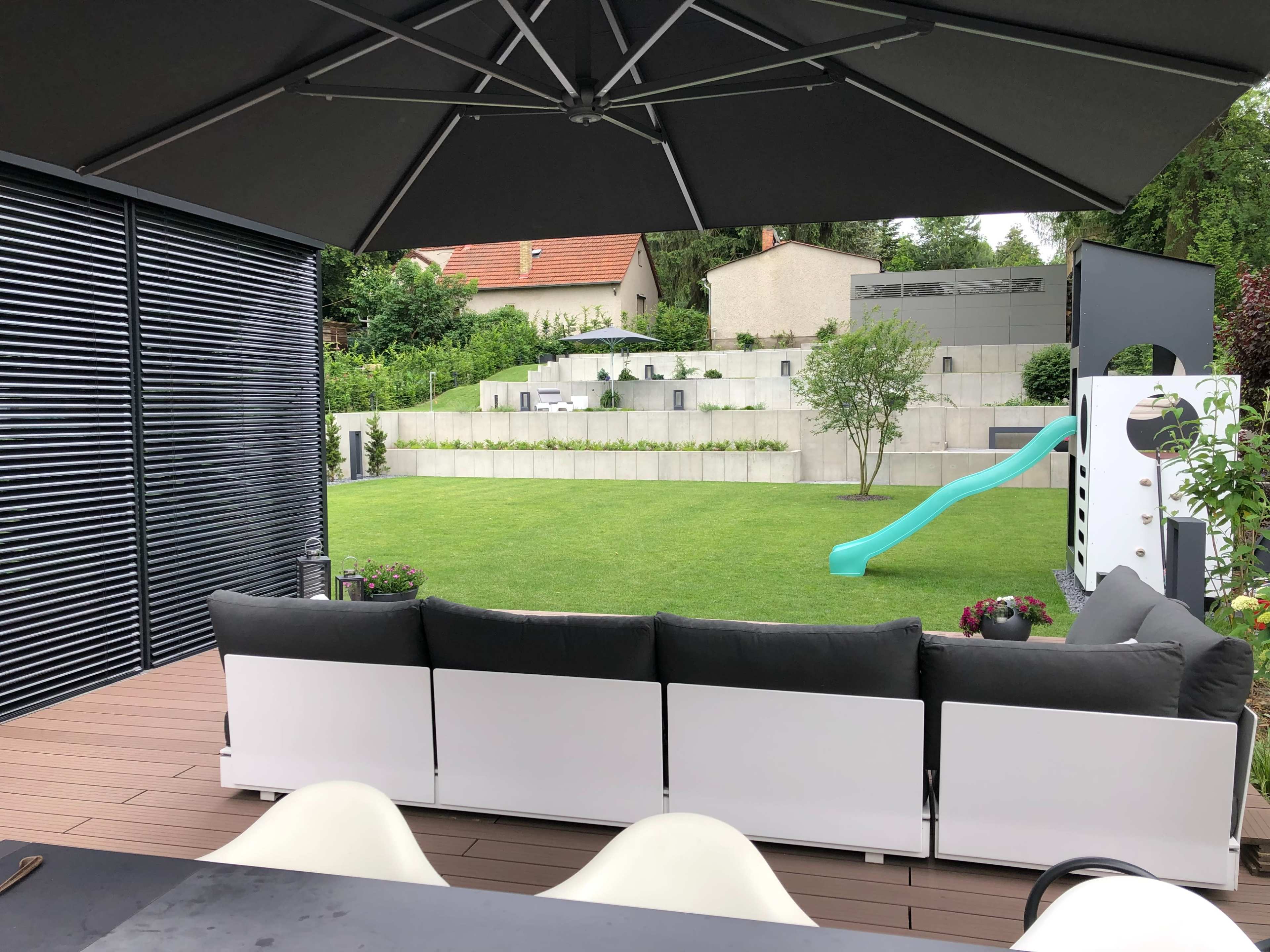 A spacious backyard with a green lawn, a slide, and a terraced area with shrubbery, viewed from a covered patio furnished with a sectional sofa.