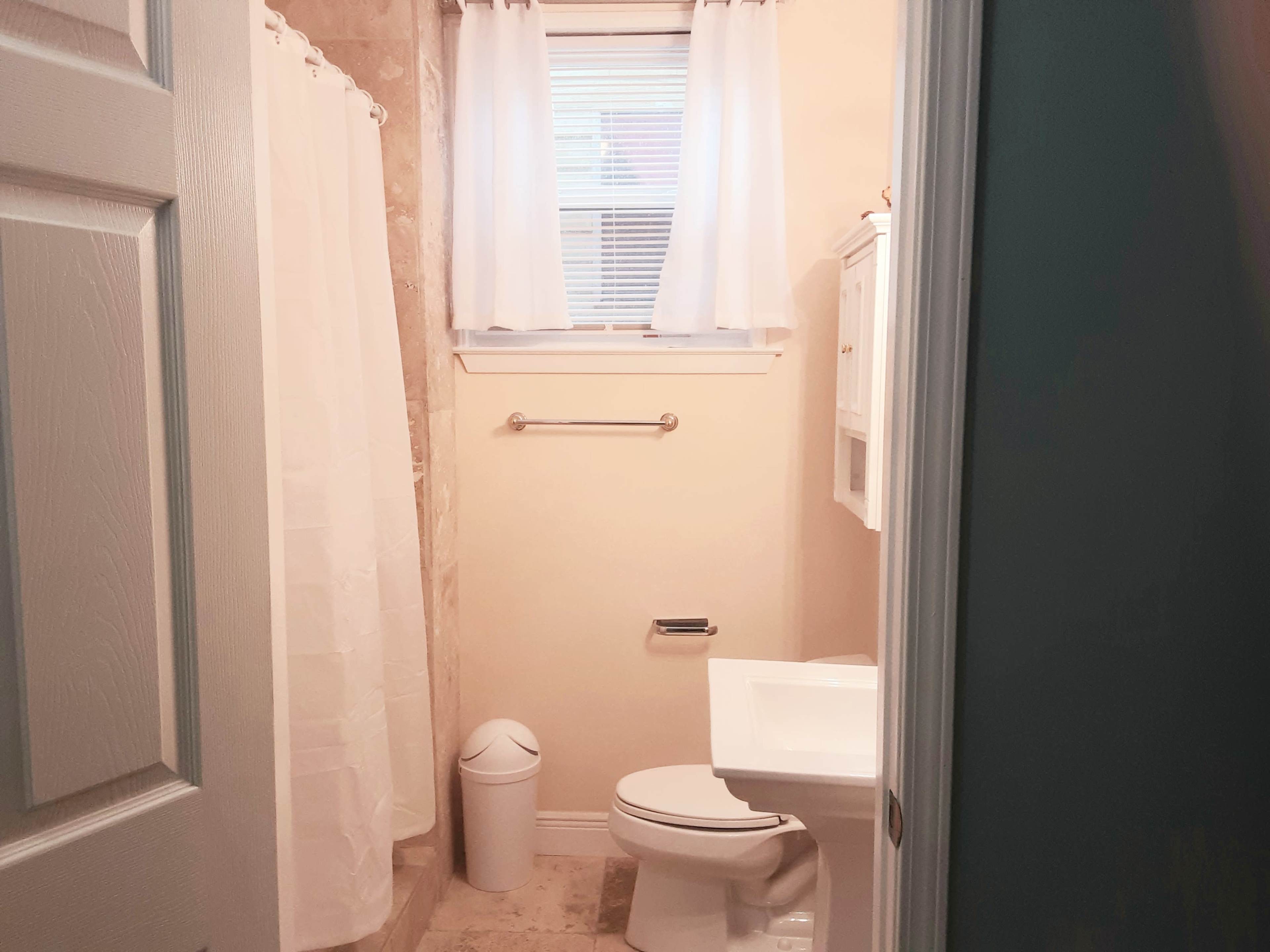 A small bathroom features a shower with a white curtain, a toilet, and a window with light curtains.