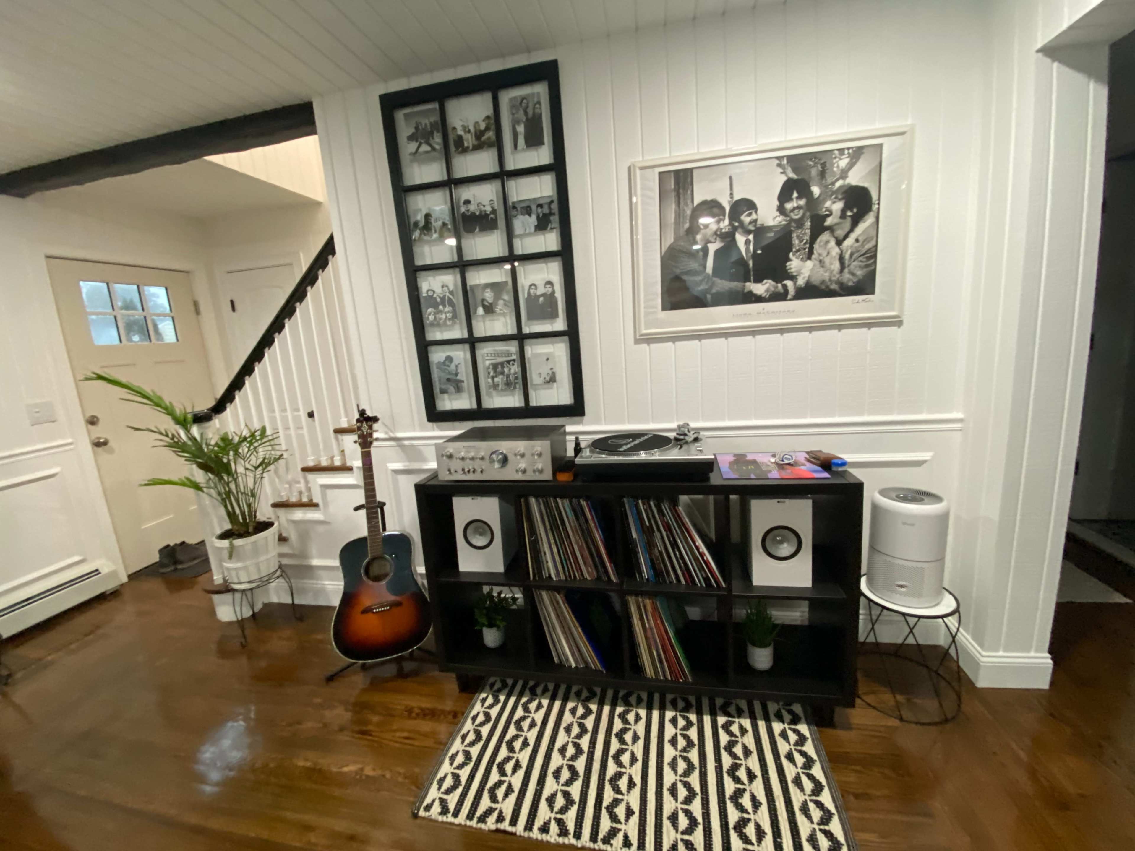The image shows a living space with a black shelving unit holding vinyl records, an acoustic guitar, and a turntable, adjacent to a staircase and framed black-and-white photos on the wall.