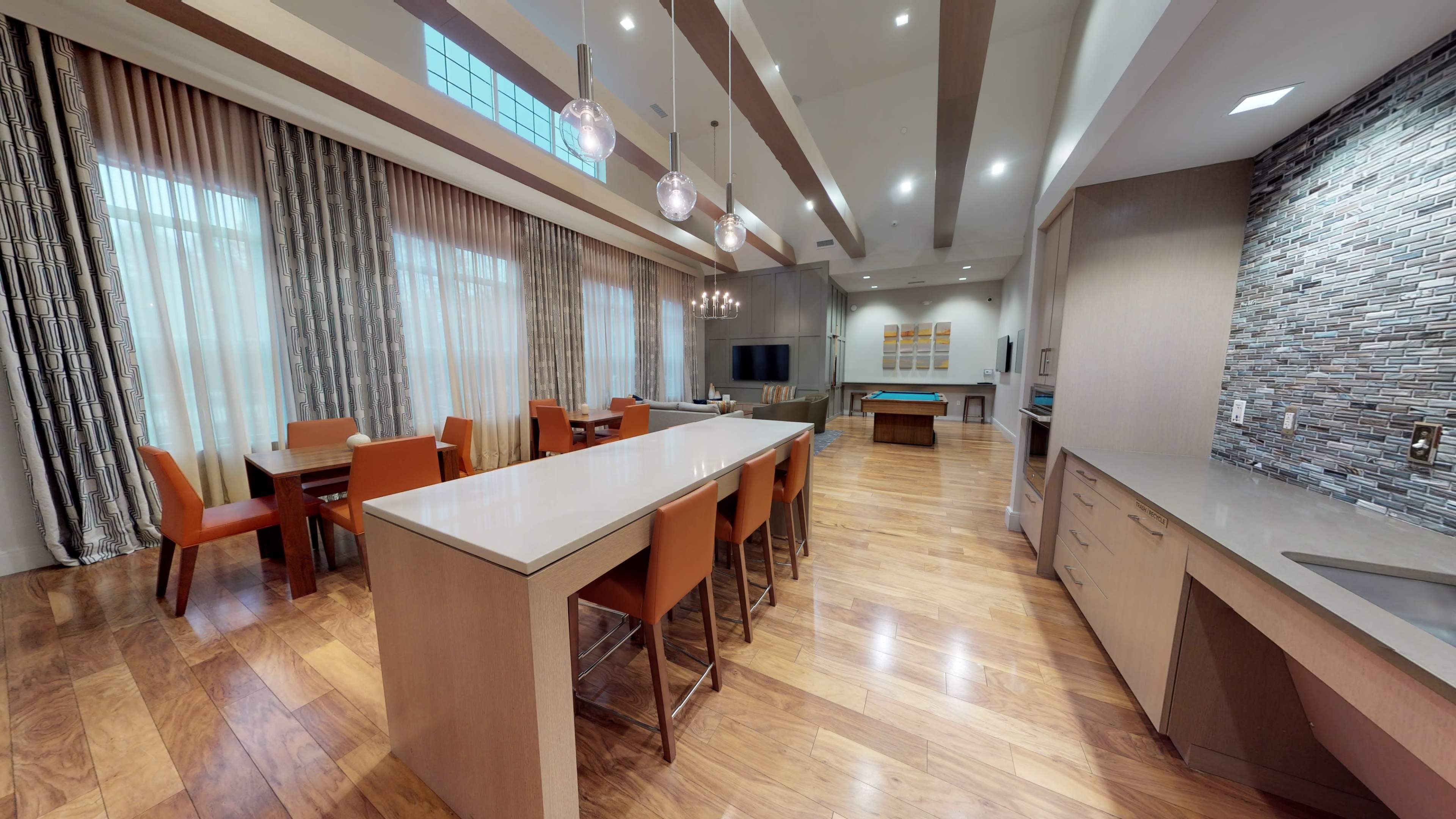 A modern dining area with a long central island, surrounded by orange chairs, leading to a spacious living area with wooden flooring and large windows.