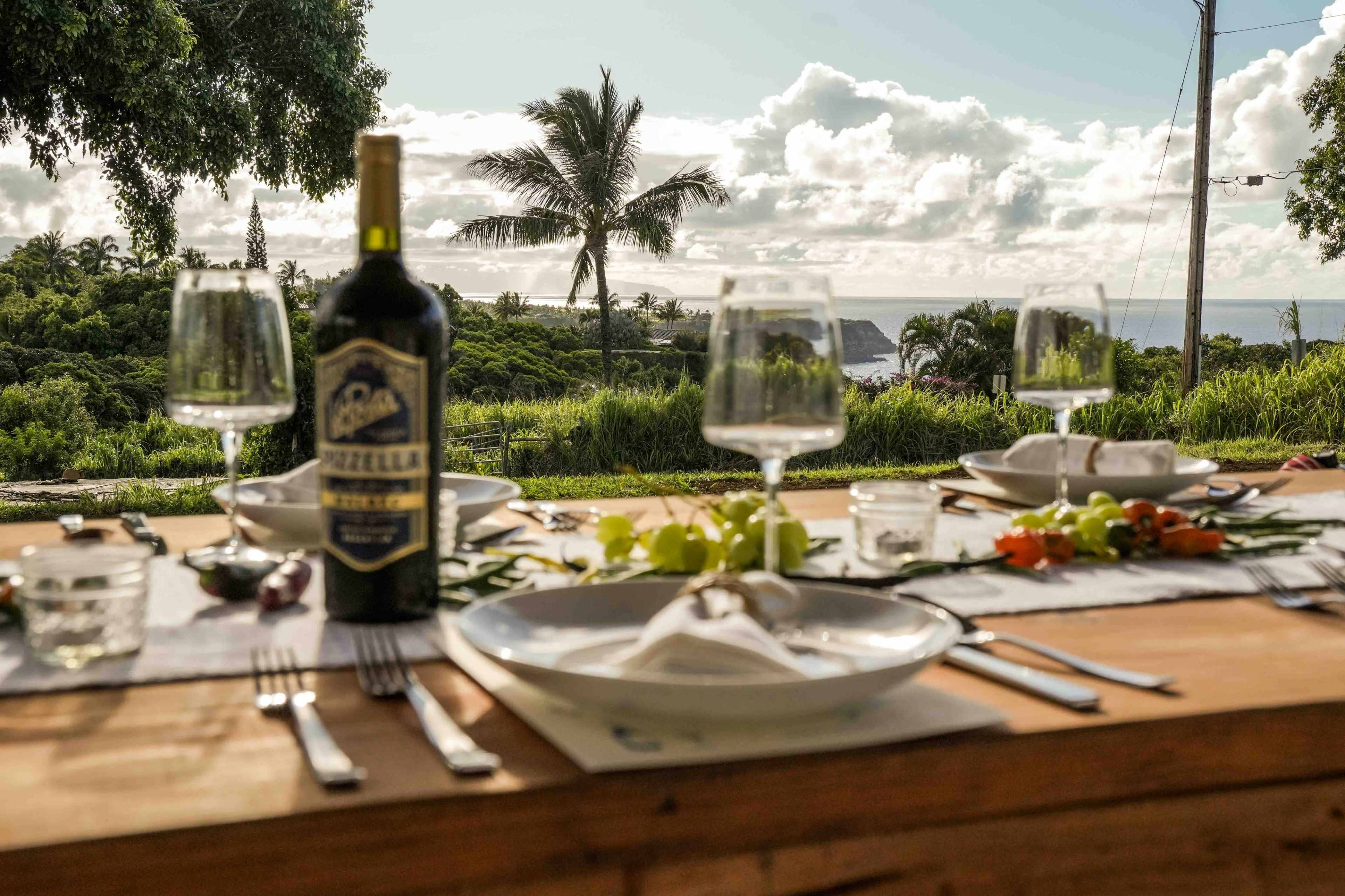 A wooden table is set for a meal with a bottle of wine, glasses, and a scenic view of palm trees and the ocean in the background.