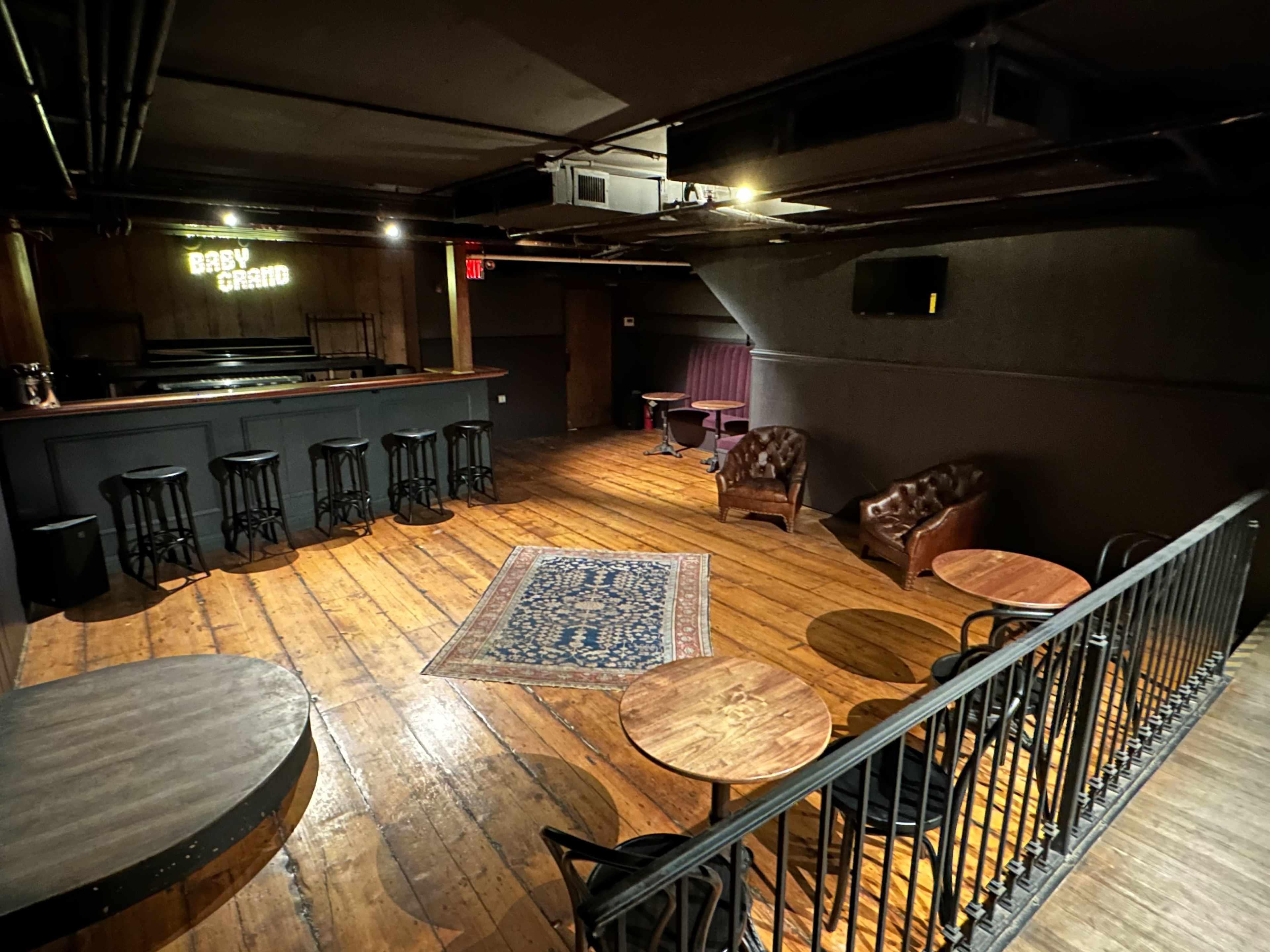 The image shows a dimly lit lounge area with wooden floors, a small stage, and several seating arrangements including leather chairs and tables.