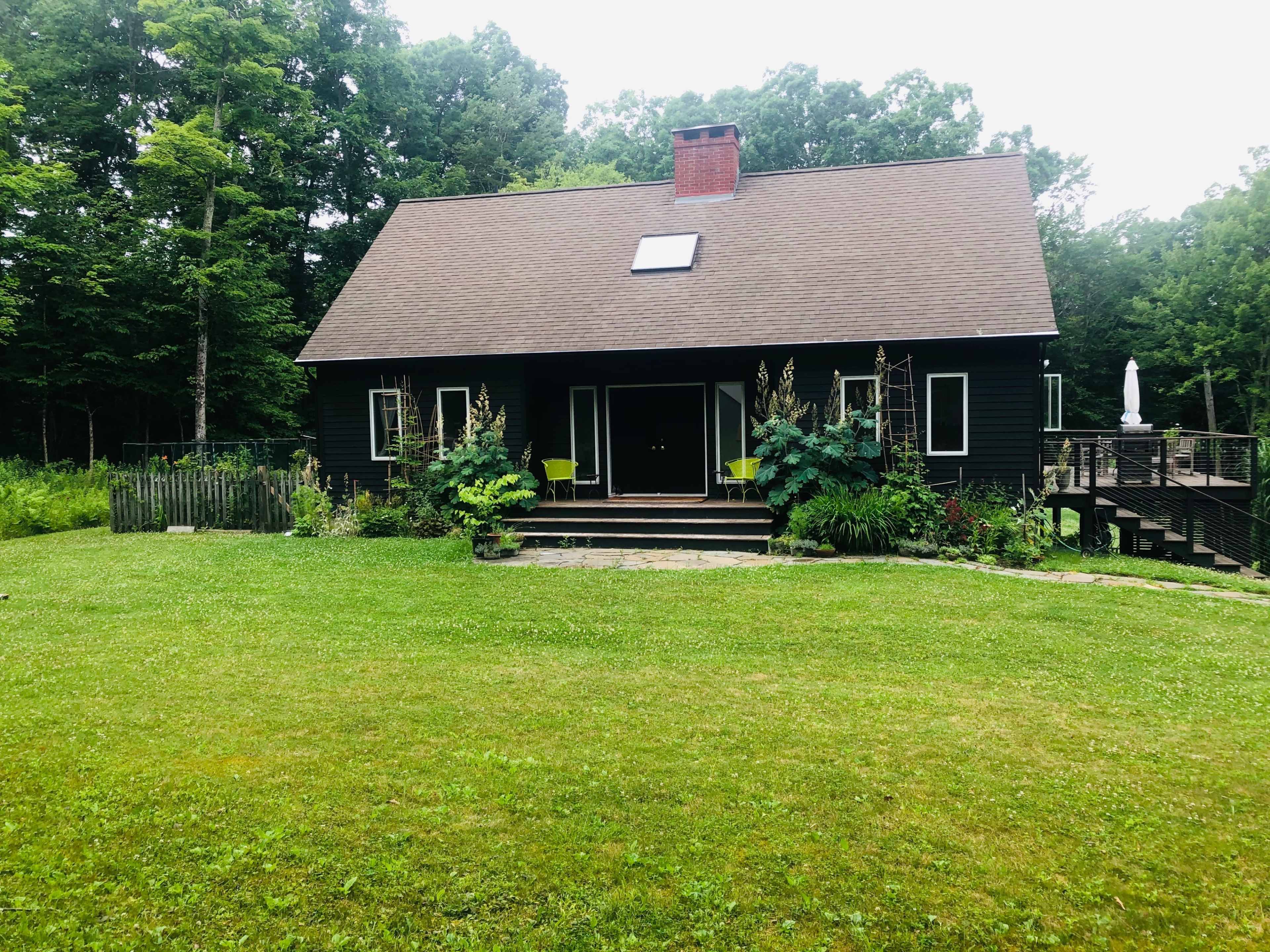 A black, two-story house with a steep roof and a front yard surrounded by trees is situated on a grassy area.