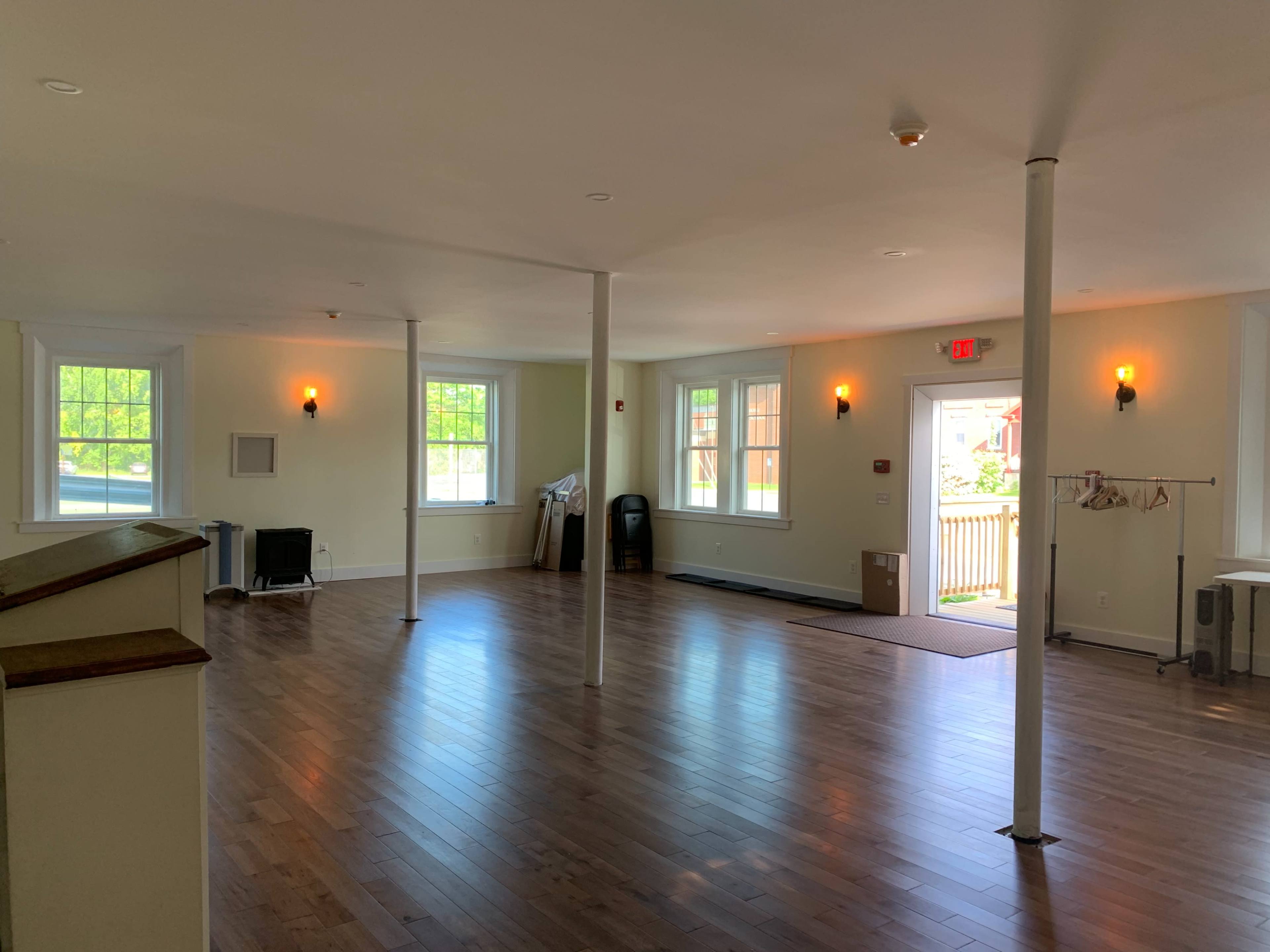 The image shows a spacious, empty room with wooden flooring, white walls, tall windows, and several columns.