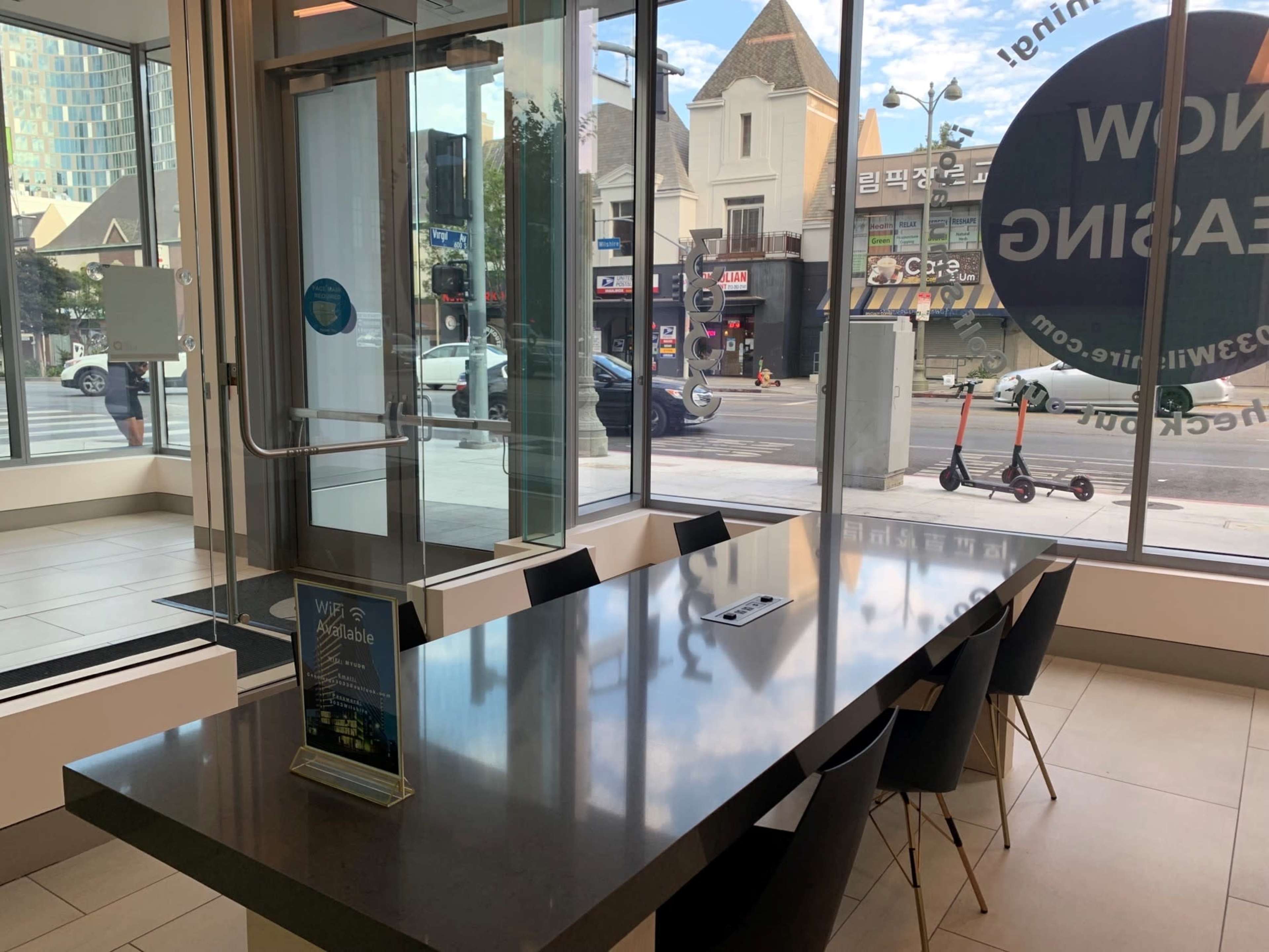 A modern storefront features a large table with chairs, facing a glass wall that overlooks a busy street.