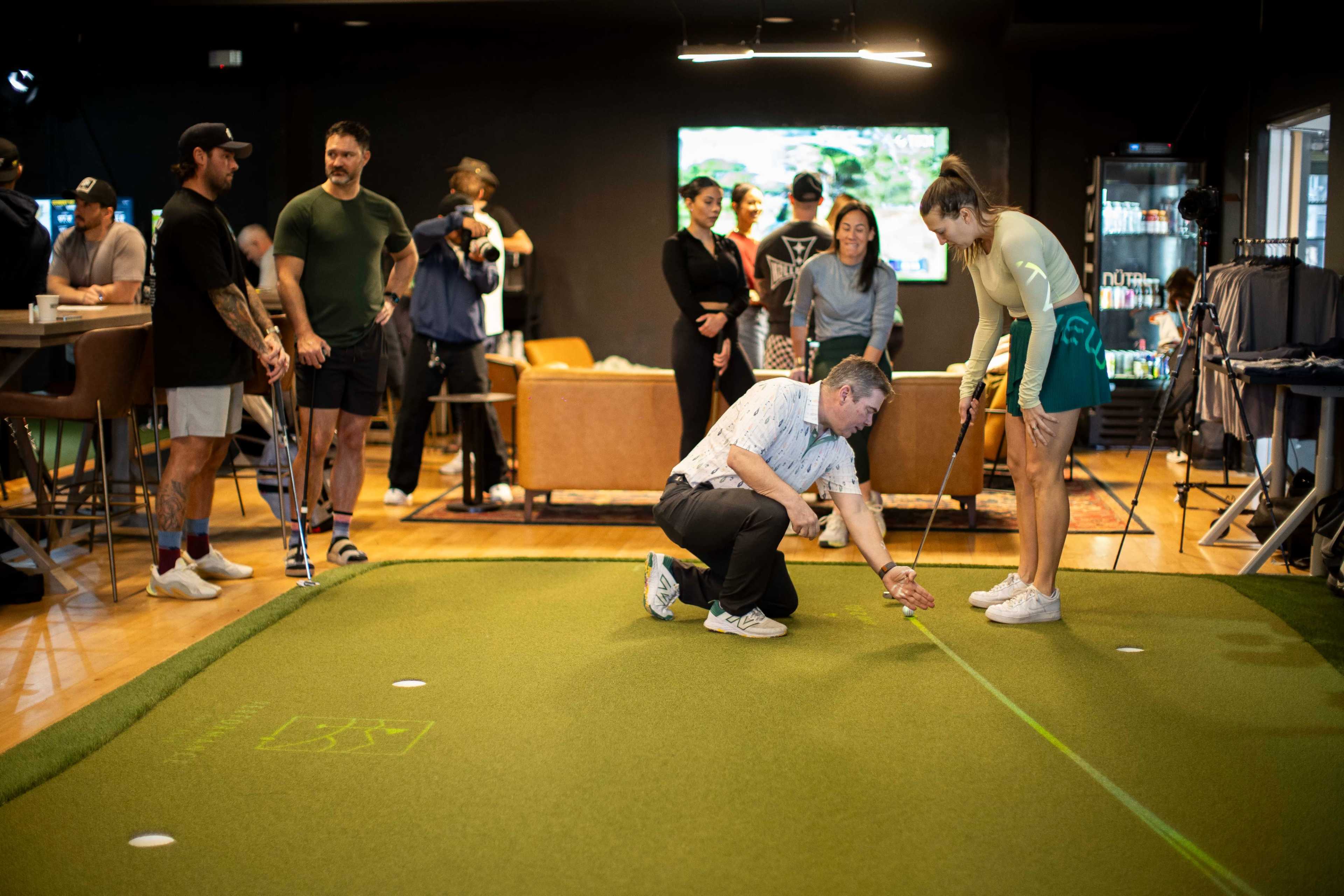 A group of people gathers around a putting green indoors, while one man demonstrates a technique to a woman.