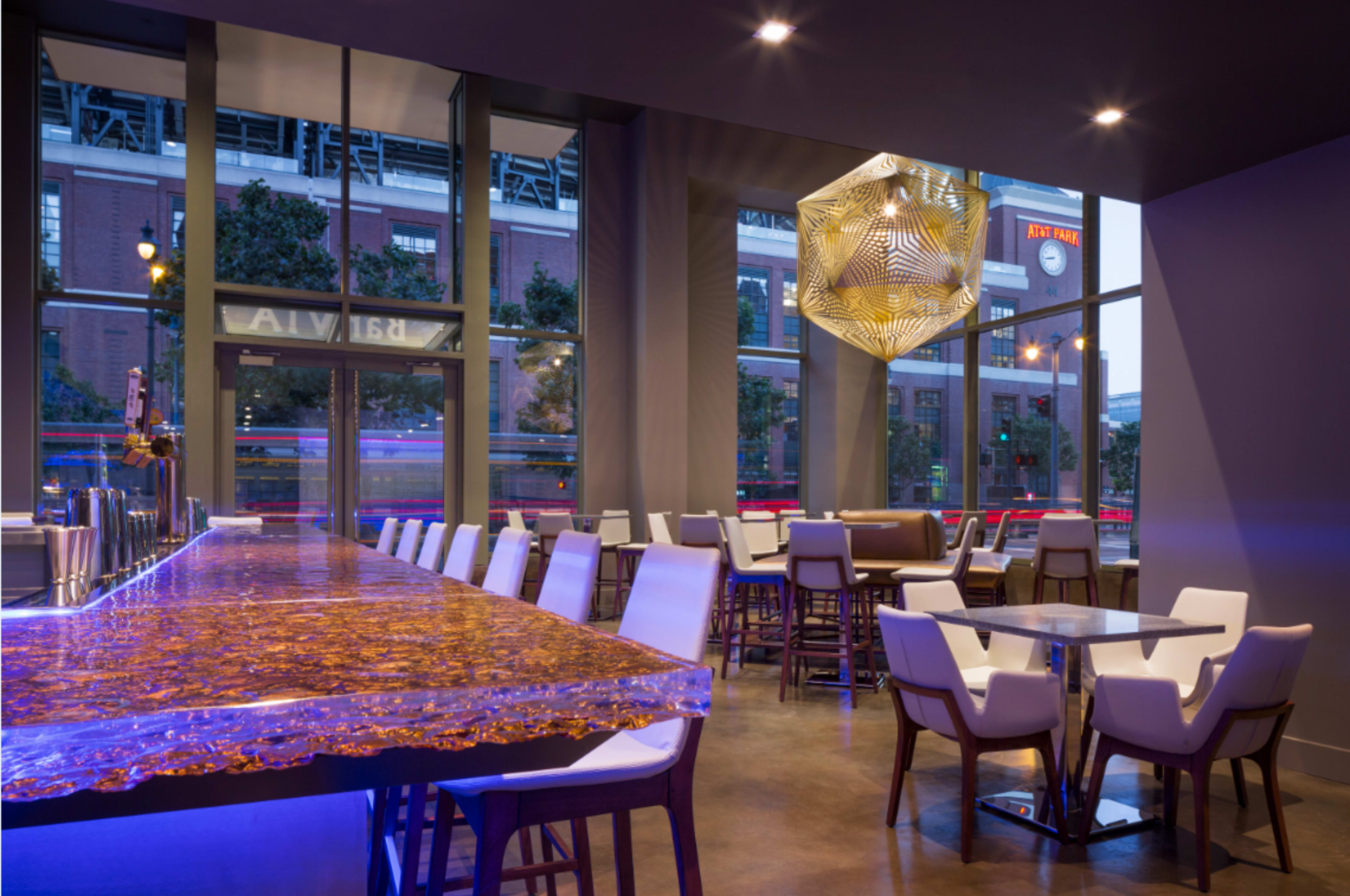 A modern interior of a bar with a glowing blue countertop, stylish chairs, and large windows revealing a view of the street outside.