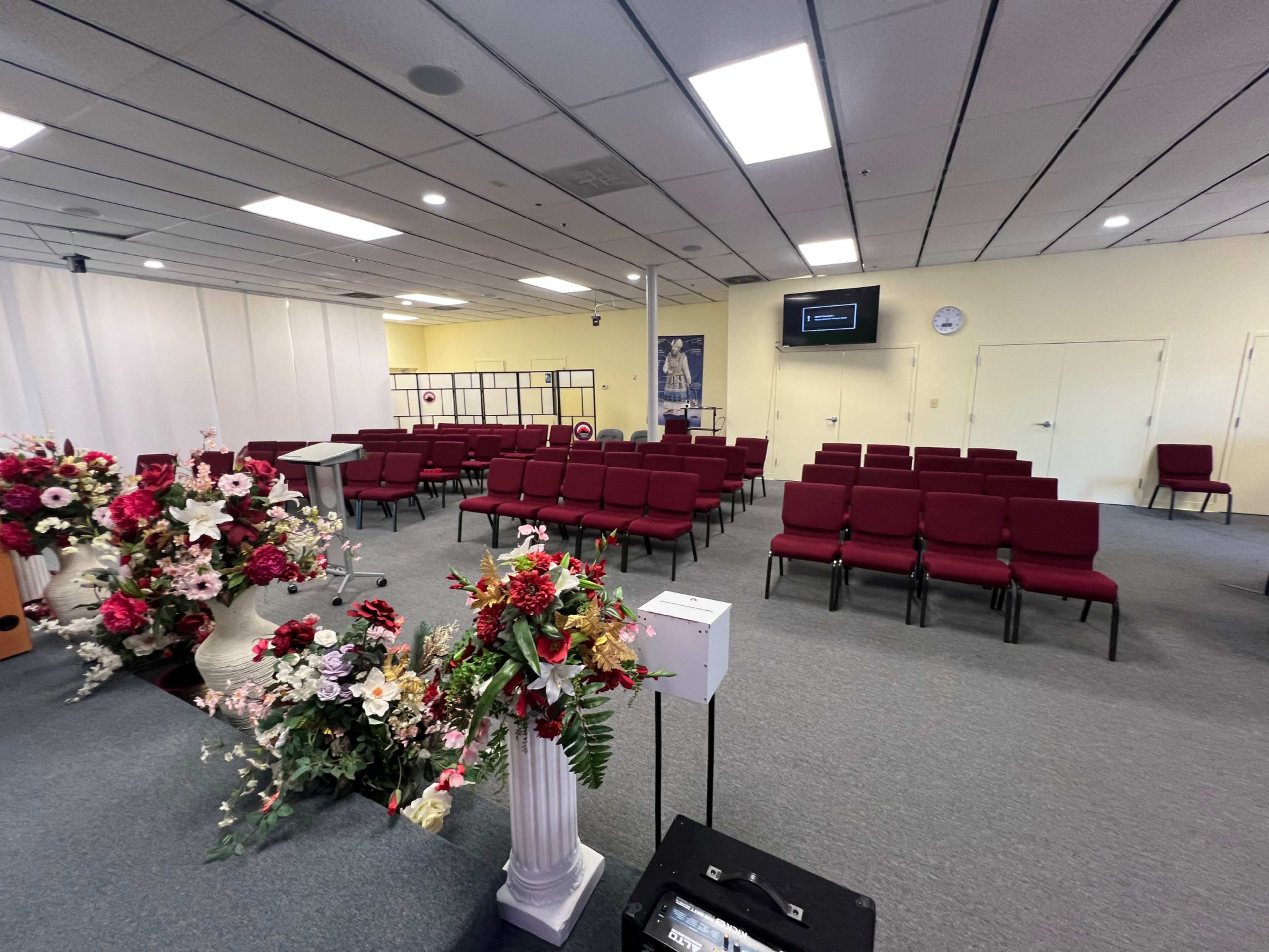 The image shows an empty church interior with rows of maroon chairs, a podium, decorative floral arrangements, and a screen displaying information.