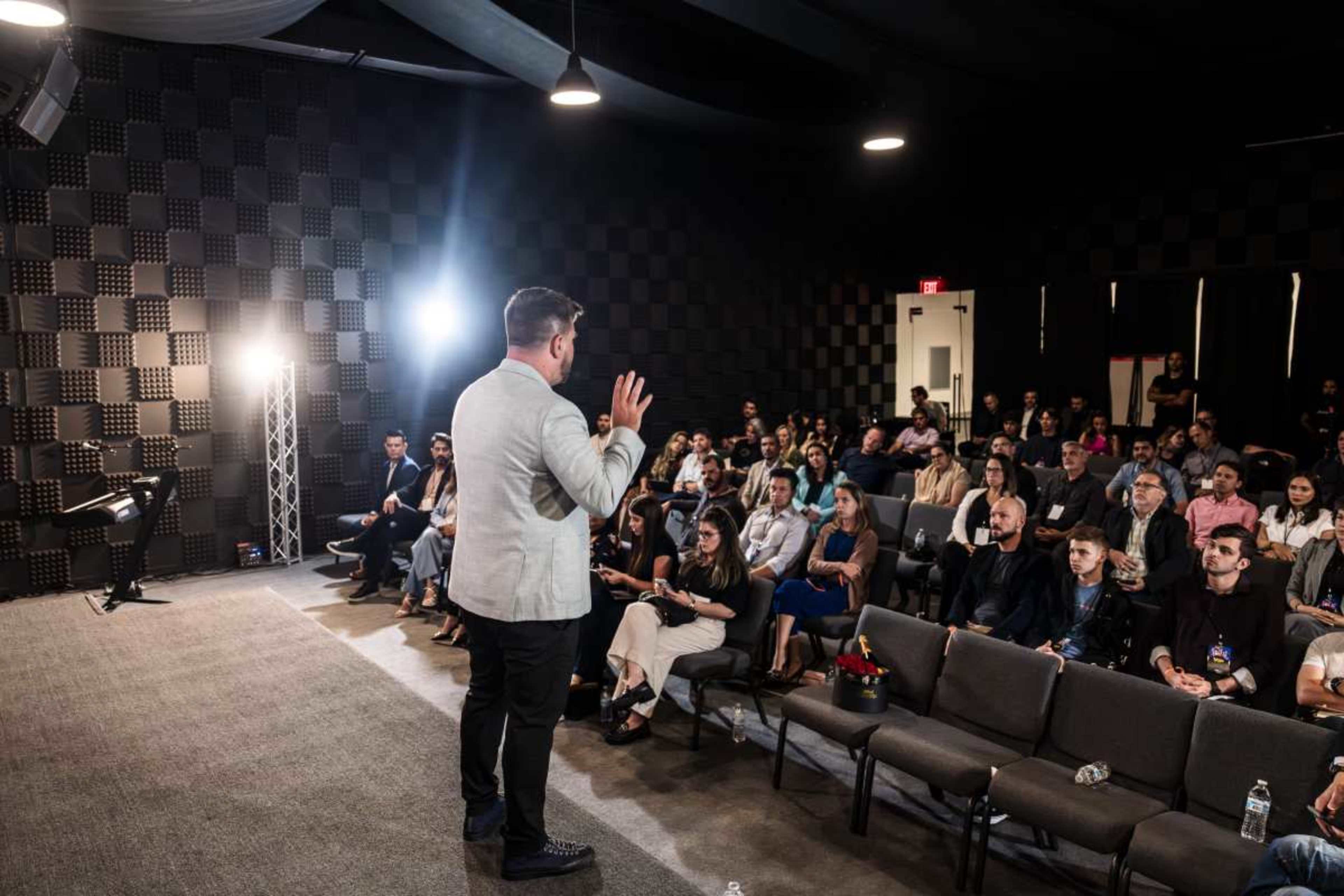 A speaker presents to an audience in a modern conference room with rows of seated attendees.