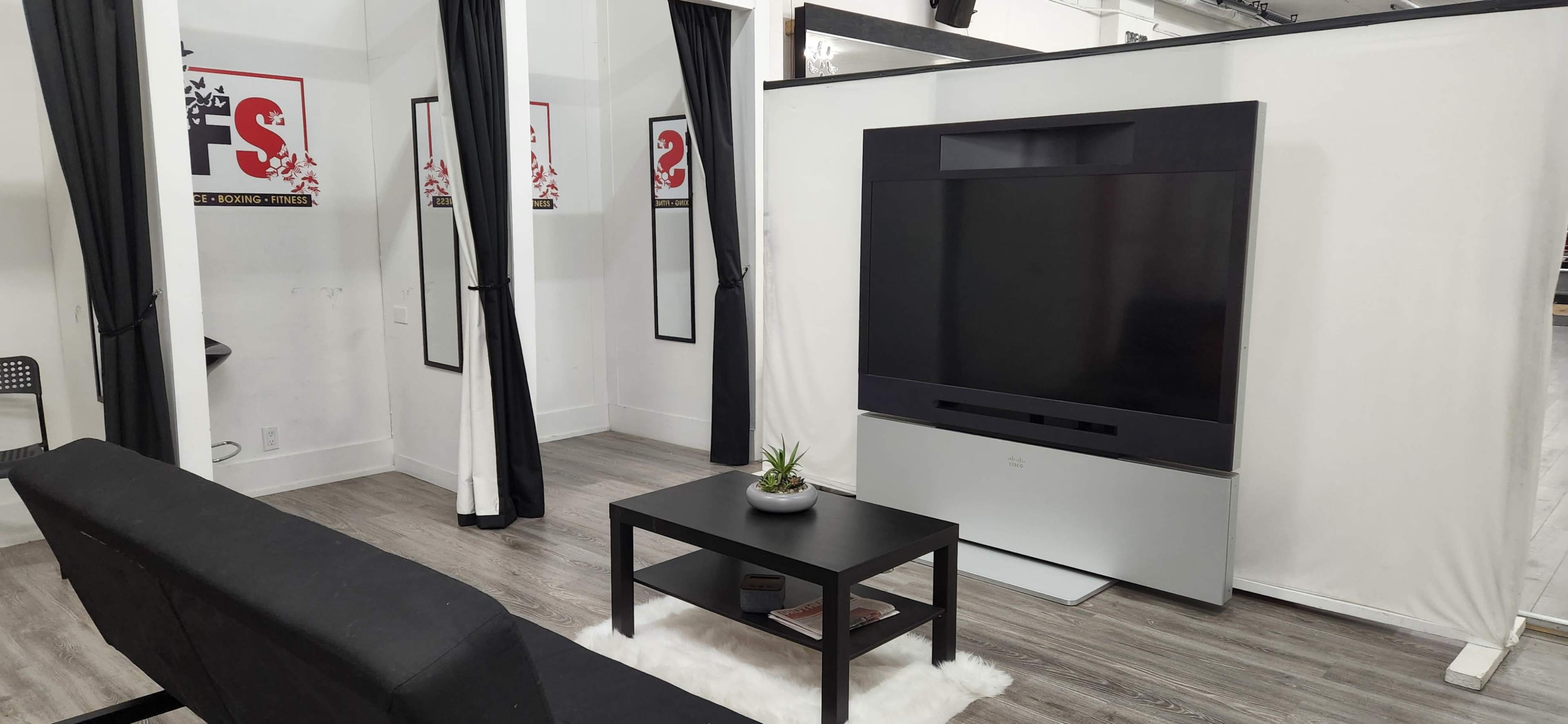 The image shows a minimalist room featuring a large television on a stand, a black coffee table, and a black sofa, with white curtains hanging in the background.