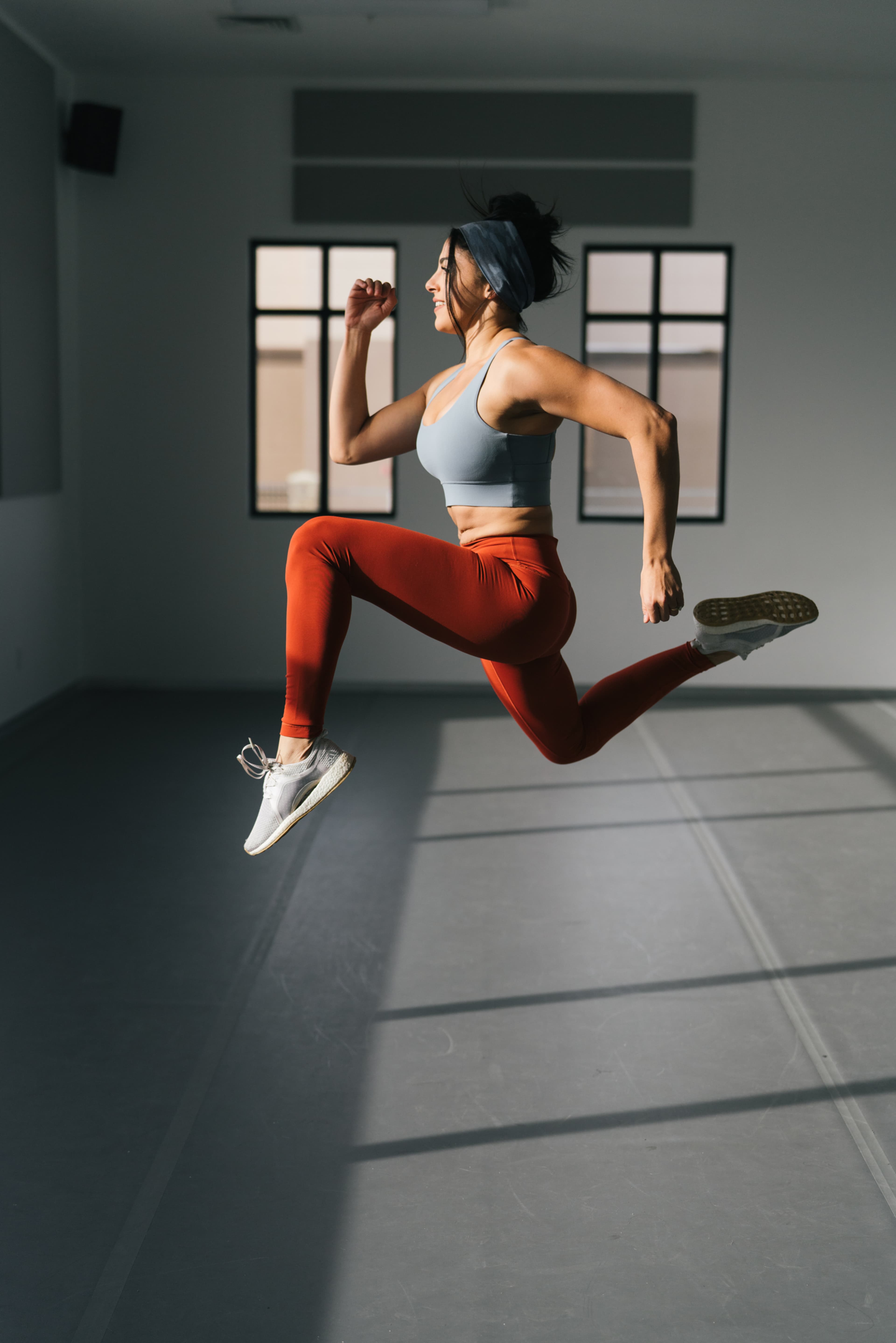 A woman is in mid-air while running, wearing a blue sports bra and orange leggings in a well-lit indoor space.