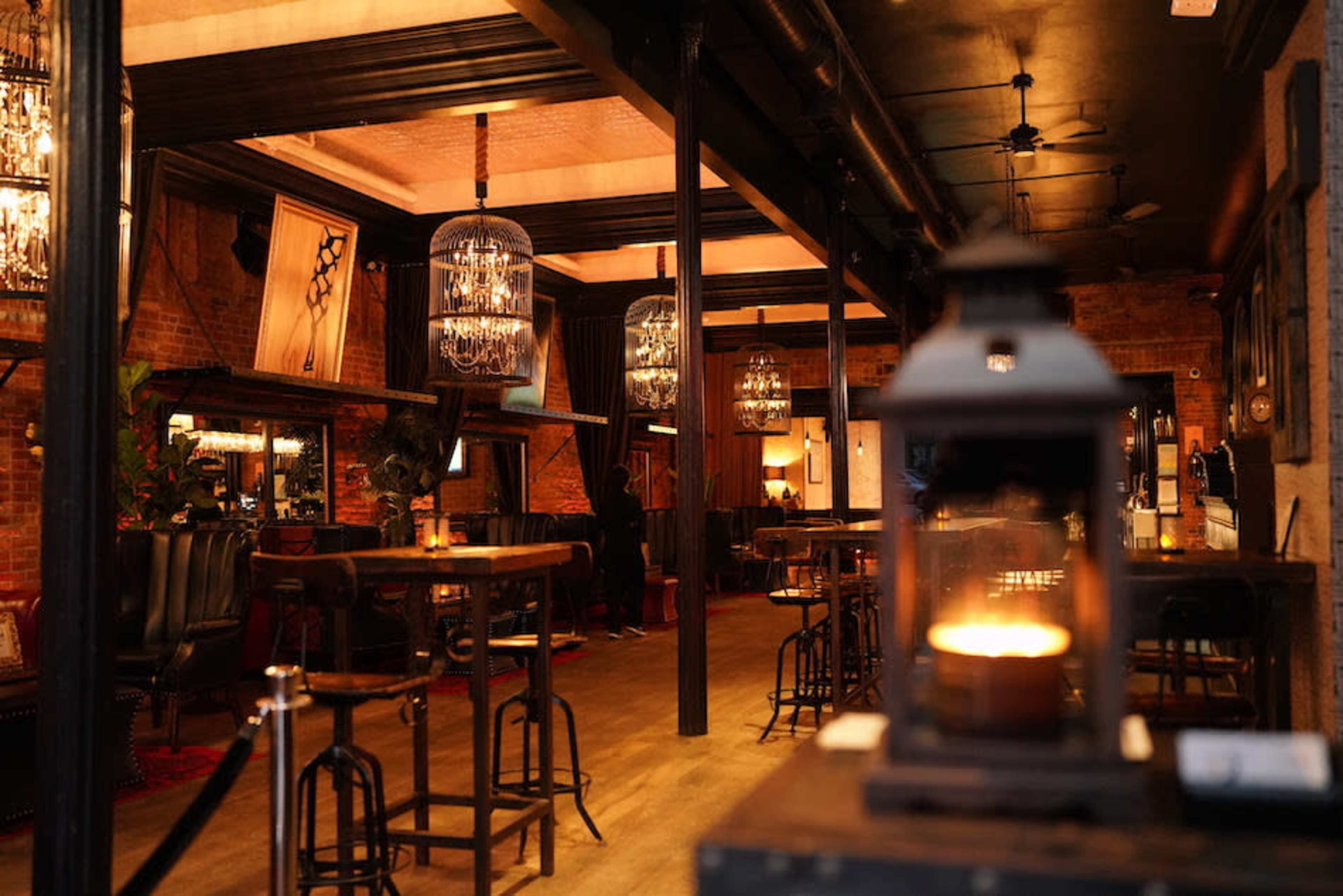 The image shows a dimly lit restaurant interior featuring wooden beams, brick walls, and ornate chandeliers hanging above bar tables and seating areas.