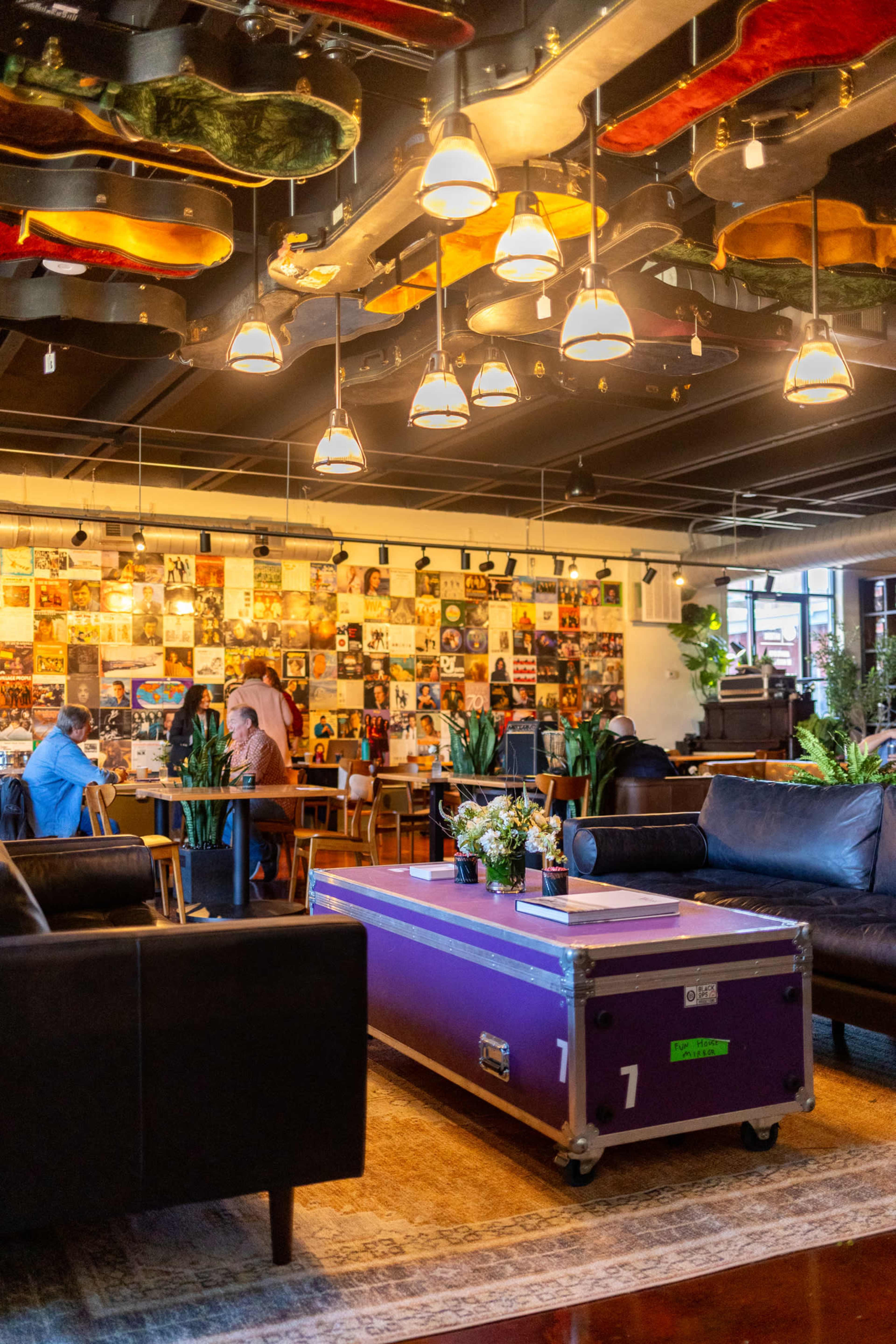The image shows a cozy lounge area with black sofas, a purple trunk, and walls decorated with framed vinyl records and artwork.