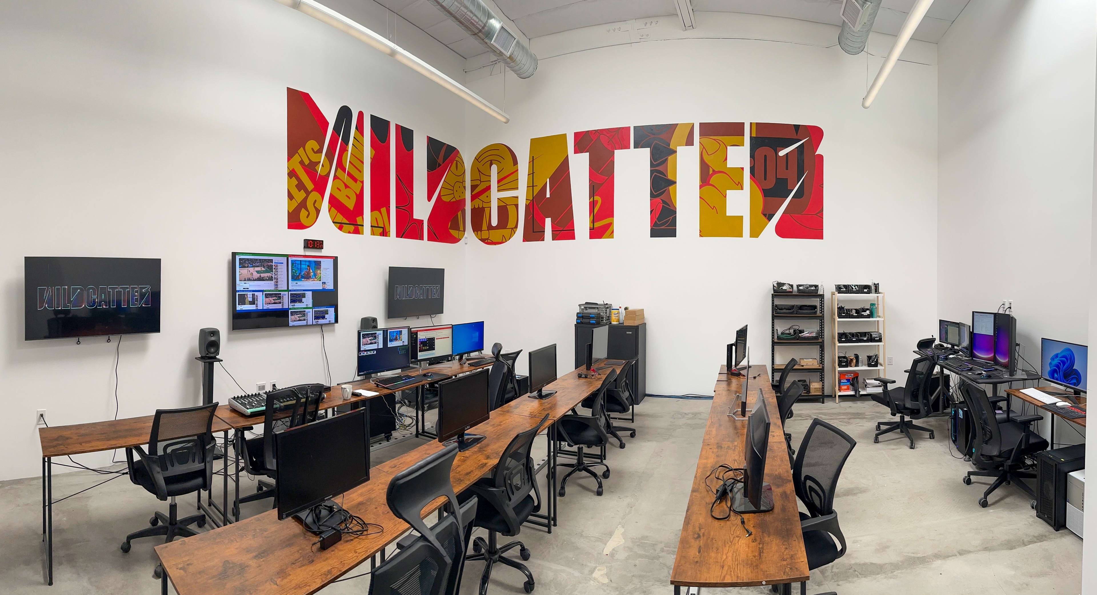 A modern workspace with several wooden tables, computers, and large monitors mounted on the walls, featuring a prominent mural that reads "Mildatter."