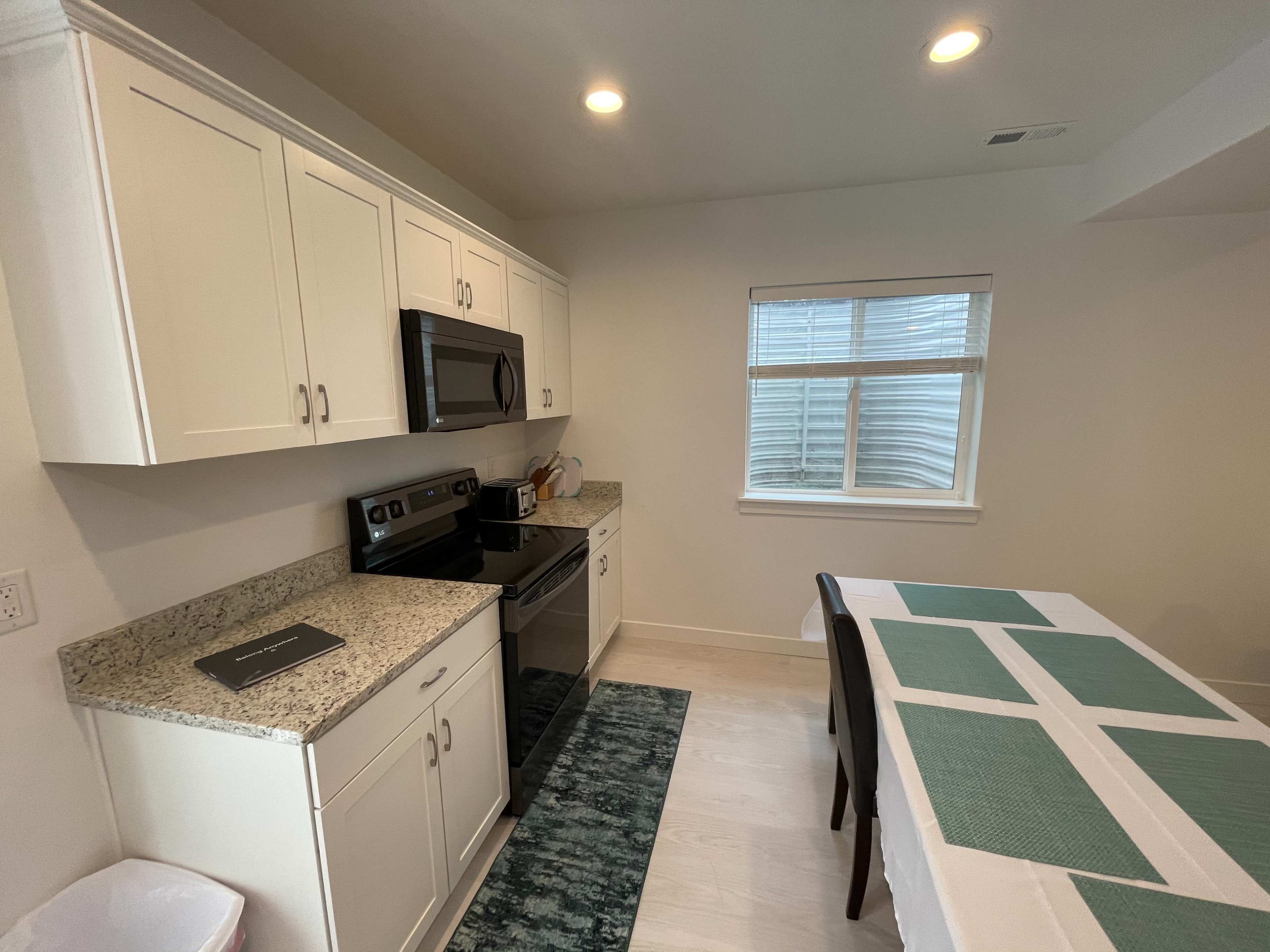 A modern kitchen with white cabinets, a stainless steel stove and microwave, and a dining area with a table covered by a green tablecloth.