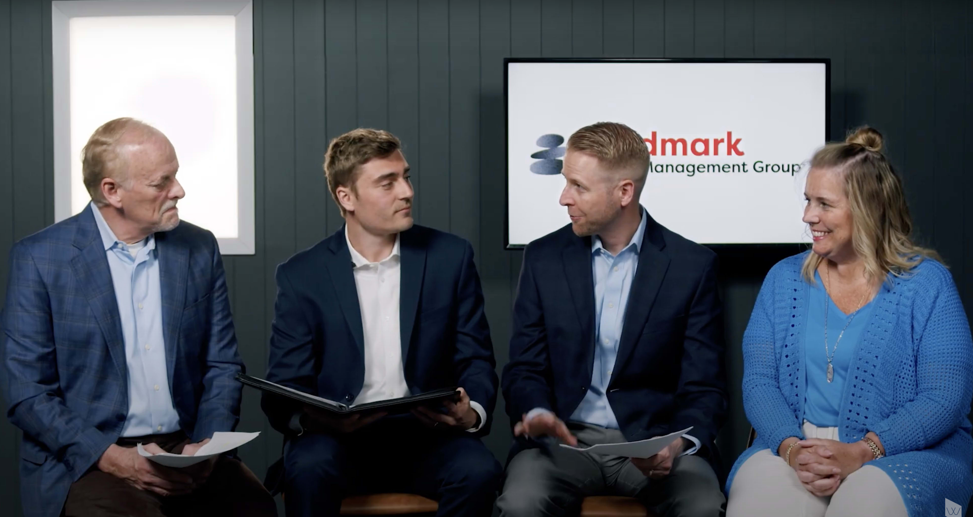 A group of four individuals sits together in front of a screen displaying the logo of Landmark Management Group, discussing with materials in hand.