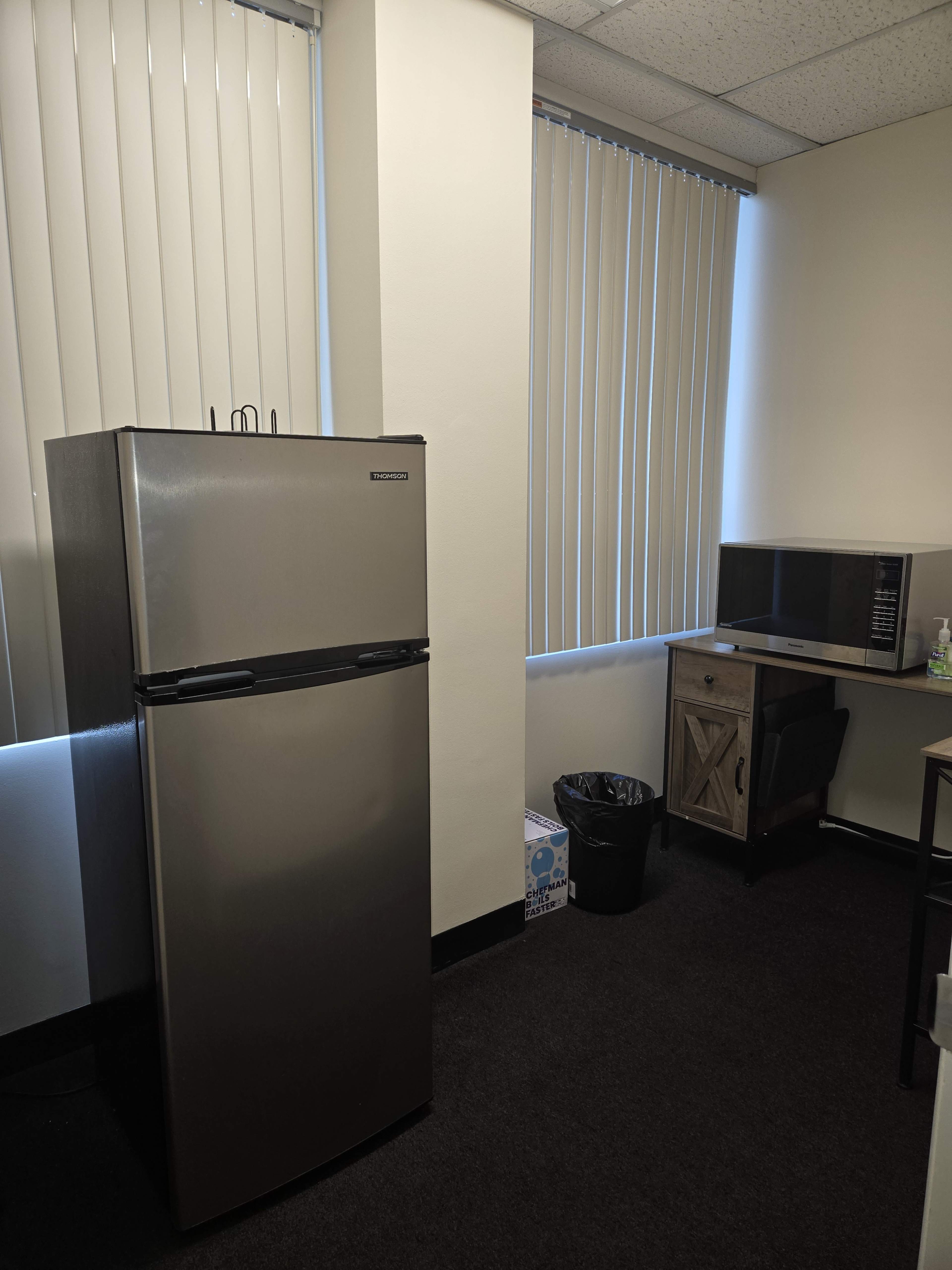 The image shows a small office space featuring a silver refrigerator, a microwave, a desk, and a trash can.