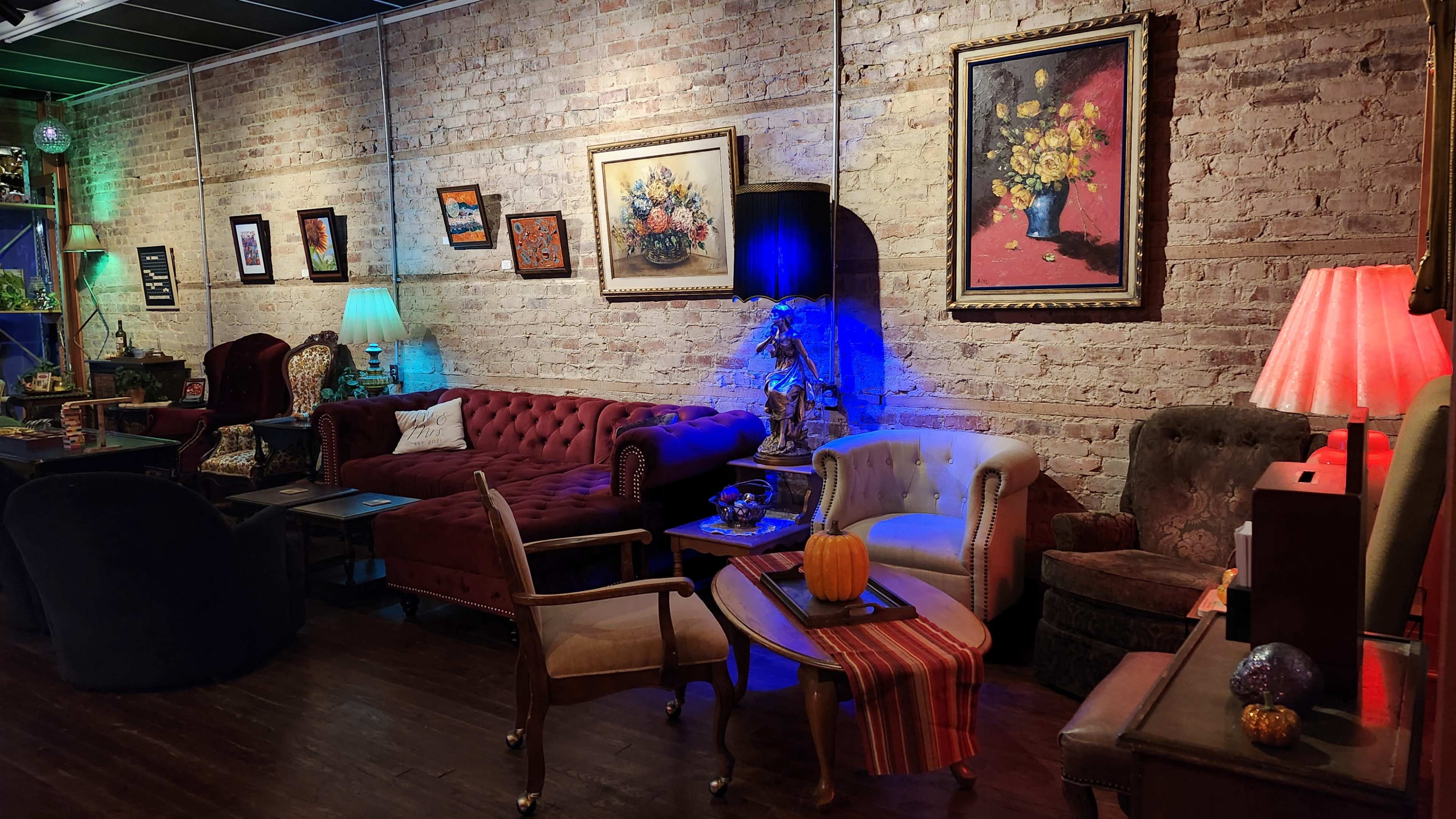 The image shows a cozy interior with various vintage-style sofas and chairs arranged against a brick wall, adorned with colorful artwork and illuminated by soft, ambient lighting.