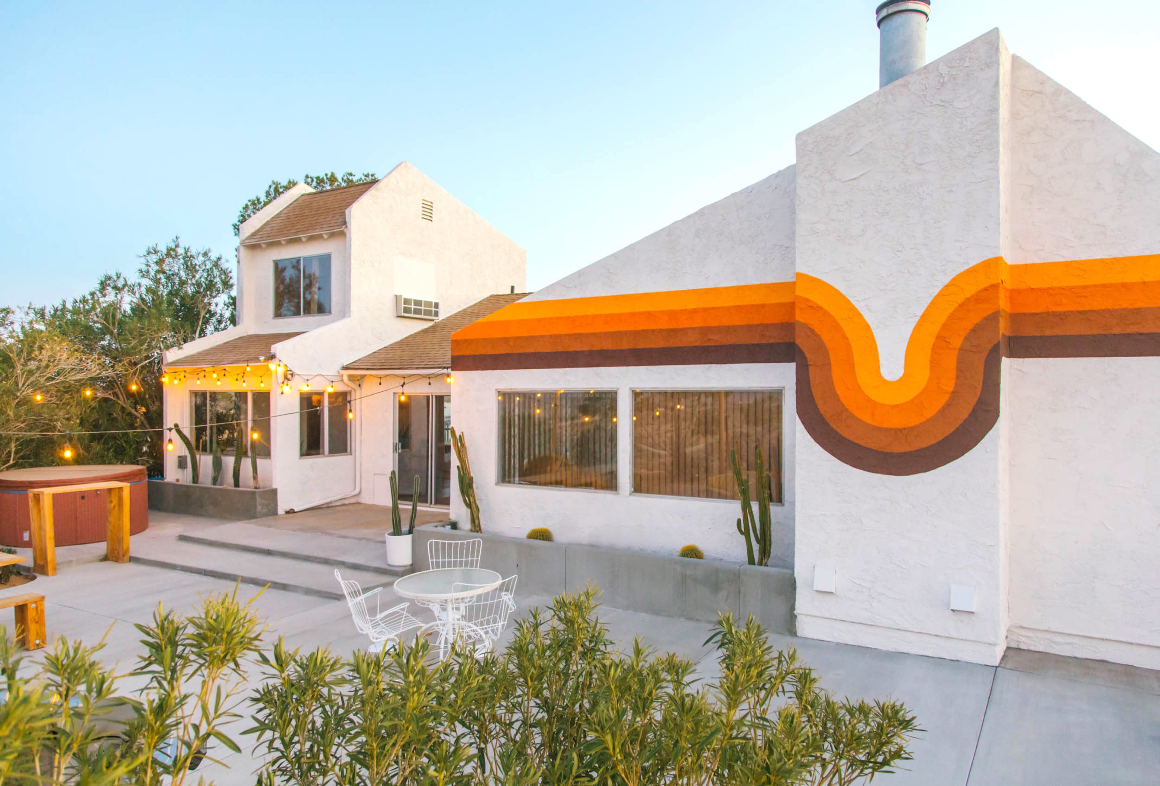 A modern two-story house features a colorful, curved mural on one side and an outdoor seating area with string lights and planters.