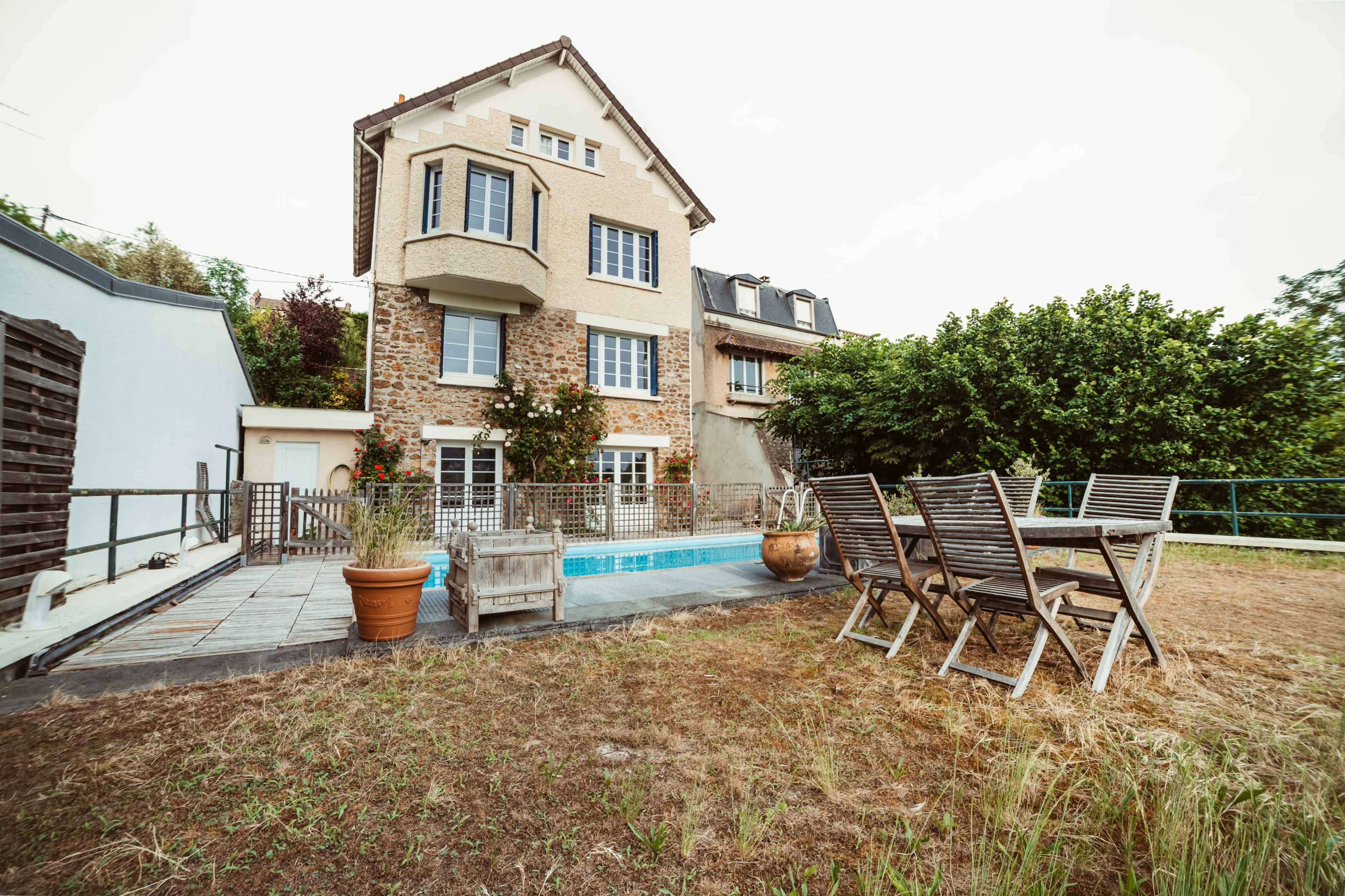 A modern house with a stone facade is surrounded by a grassy area, featuring a small swimming pool and a wooden dining set on a patio.