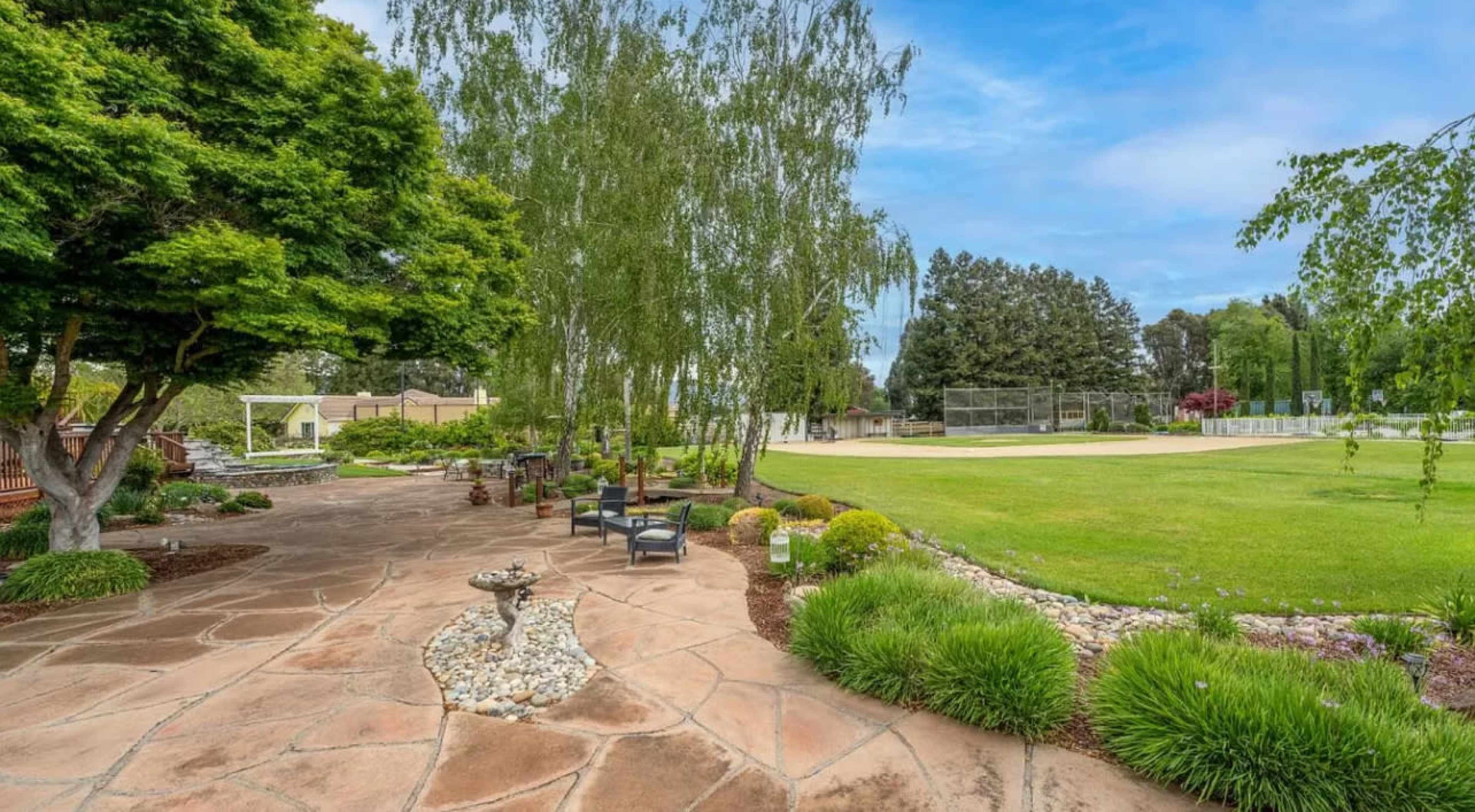 A landscaped outdoor space with stone pathways, seating areas, and a view of a grassy field and trees in the background.