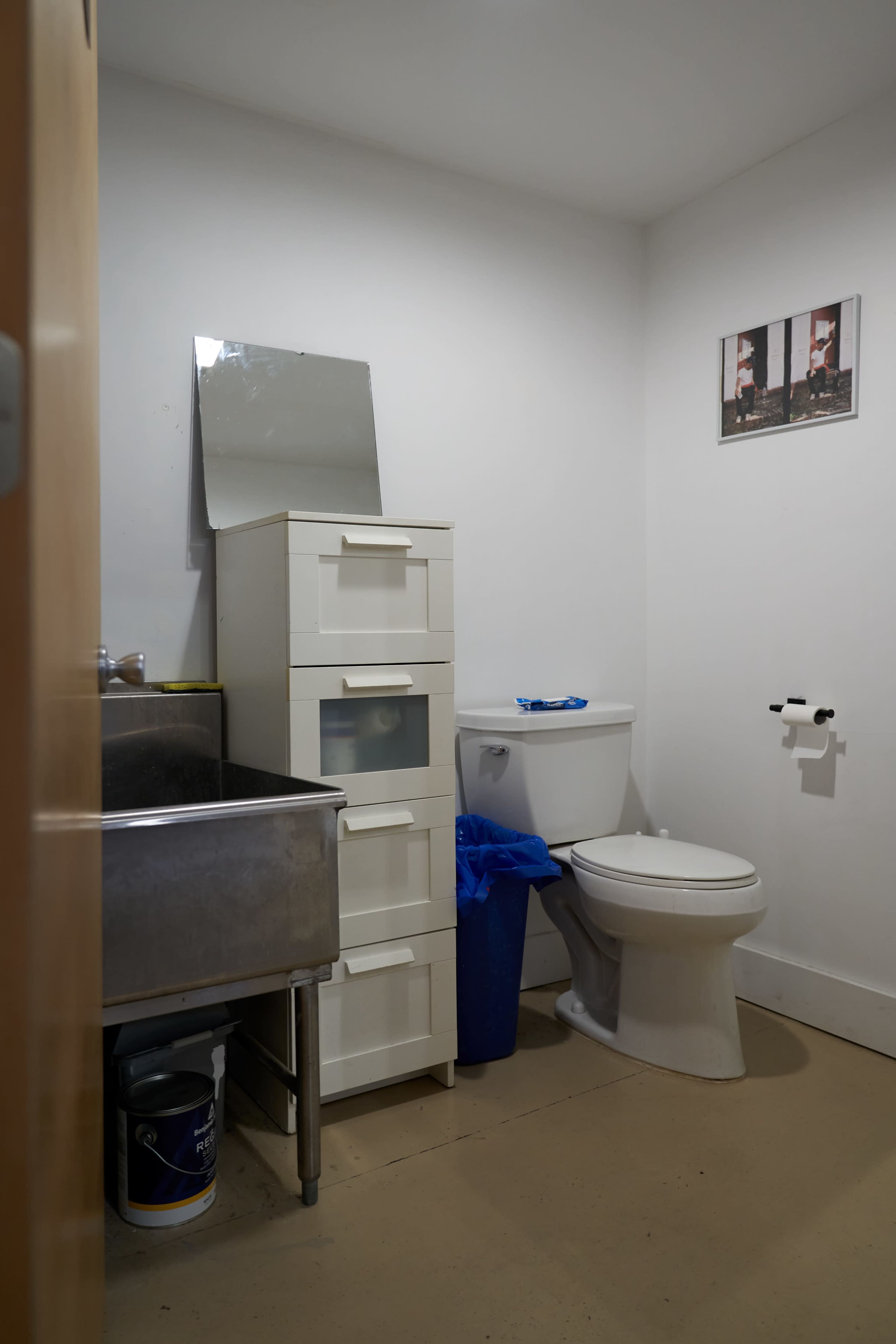 The image shows a small bathroom with a white toilet, a sink with a metal cabinet beside it, a blue trash bin, and a small mirror on the wall.