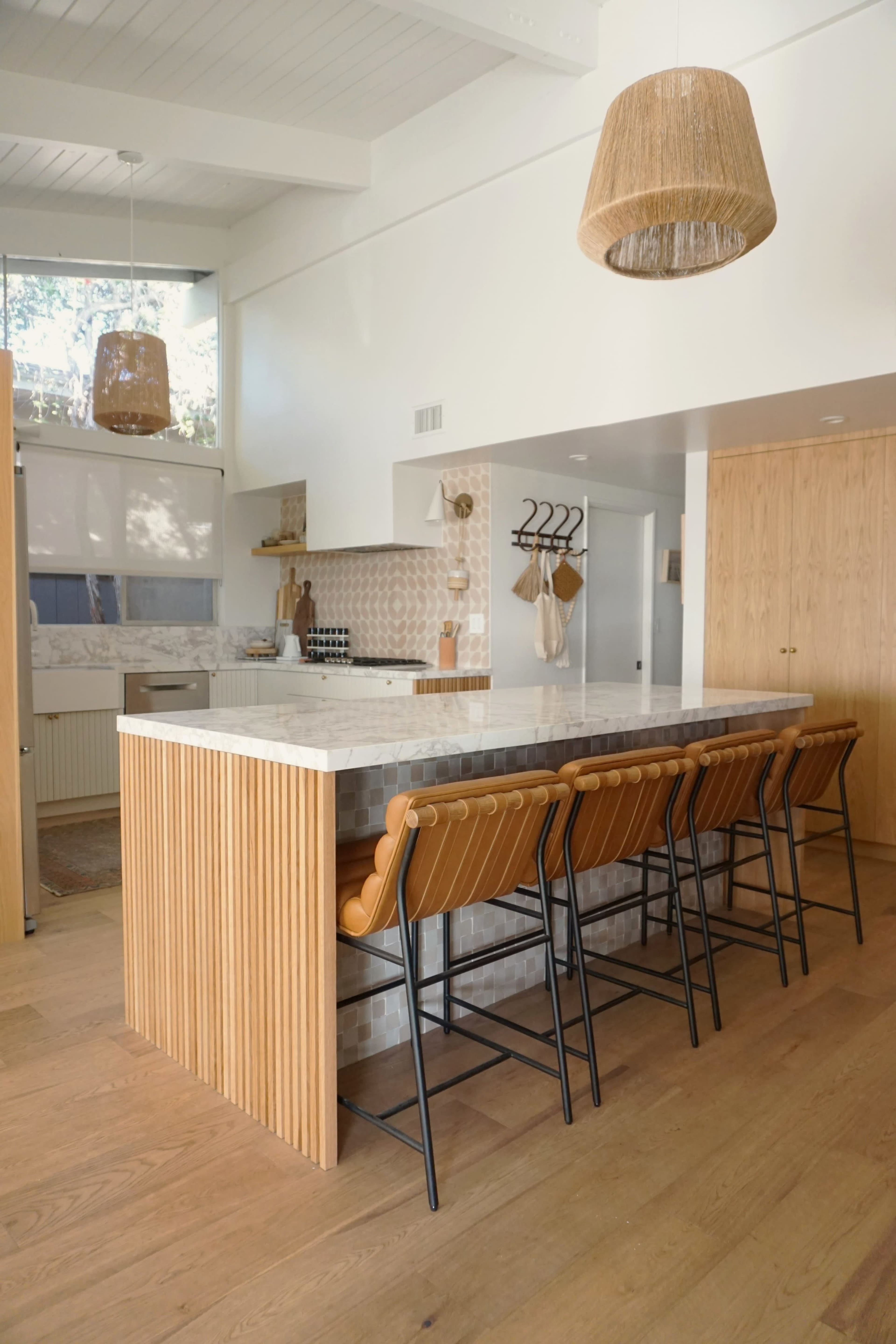 A modern kitchen features a marble island with wooden accents, surrounded by high-backed bar stools, and is illuminated by woven pendant lights.