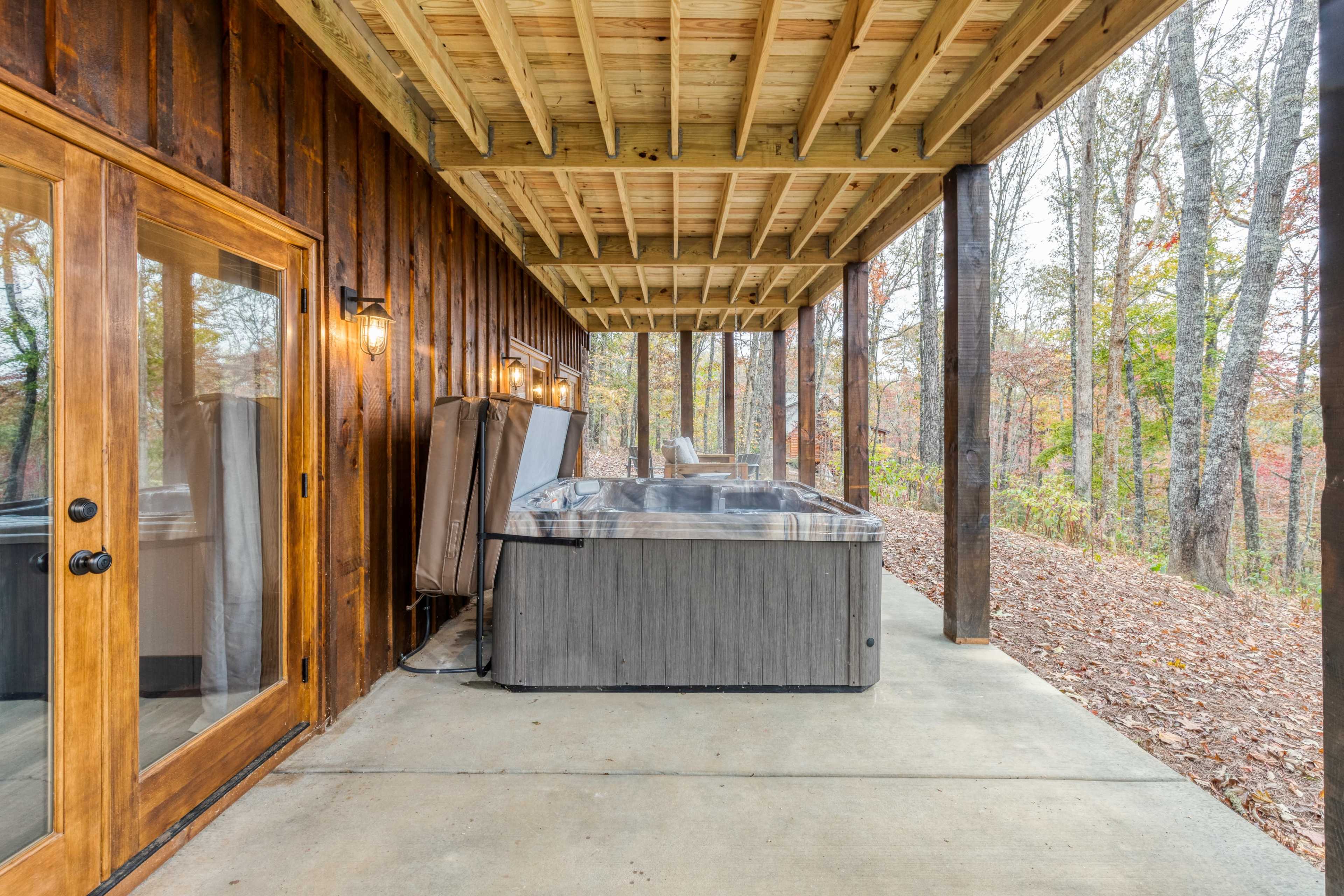 A hot tub is situated on a concrete patio beneath a wooden overhang, surrounded by trees in a wooded area.