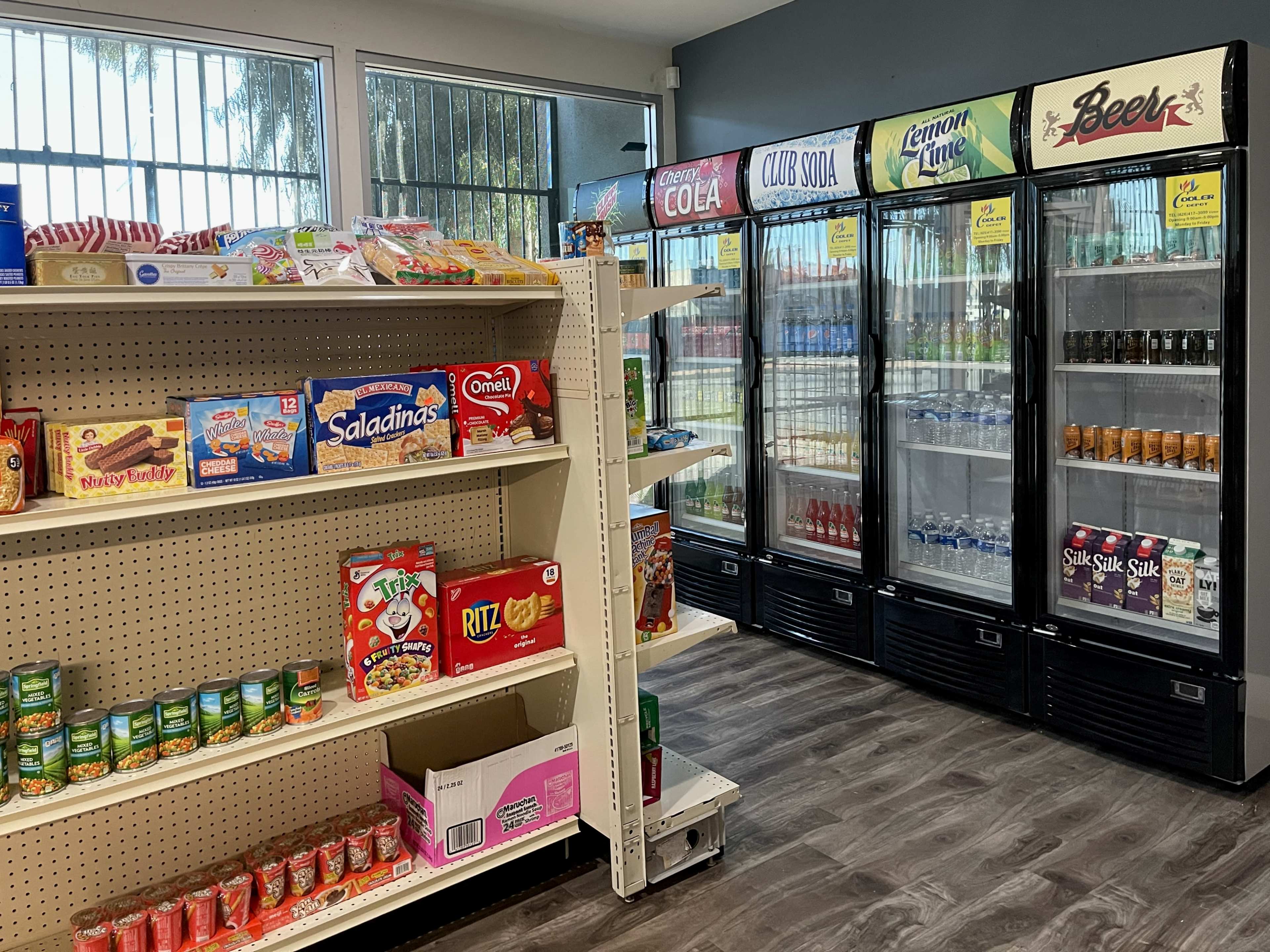 An interior of a convenience store with shelves stocked with snacks on the left and a display of refrigerated beverages on the right.