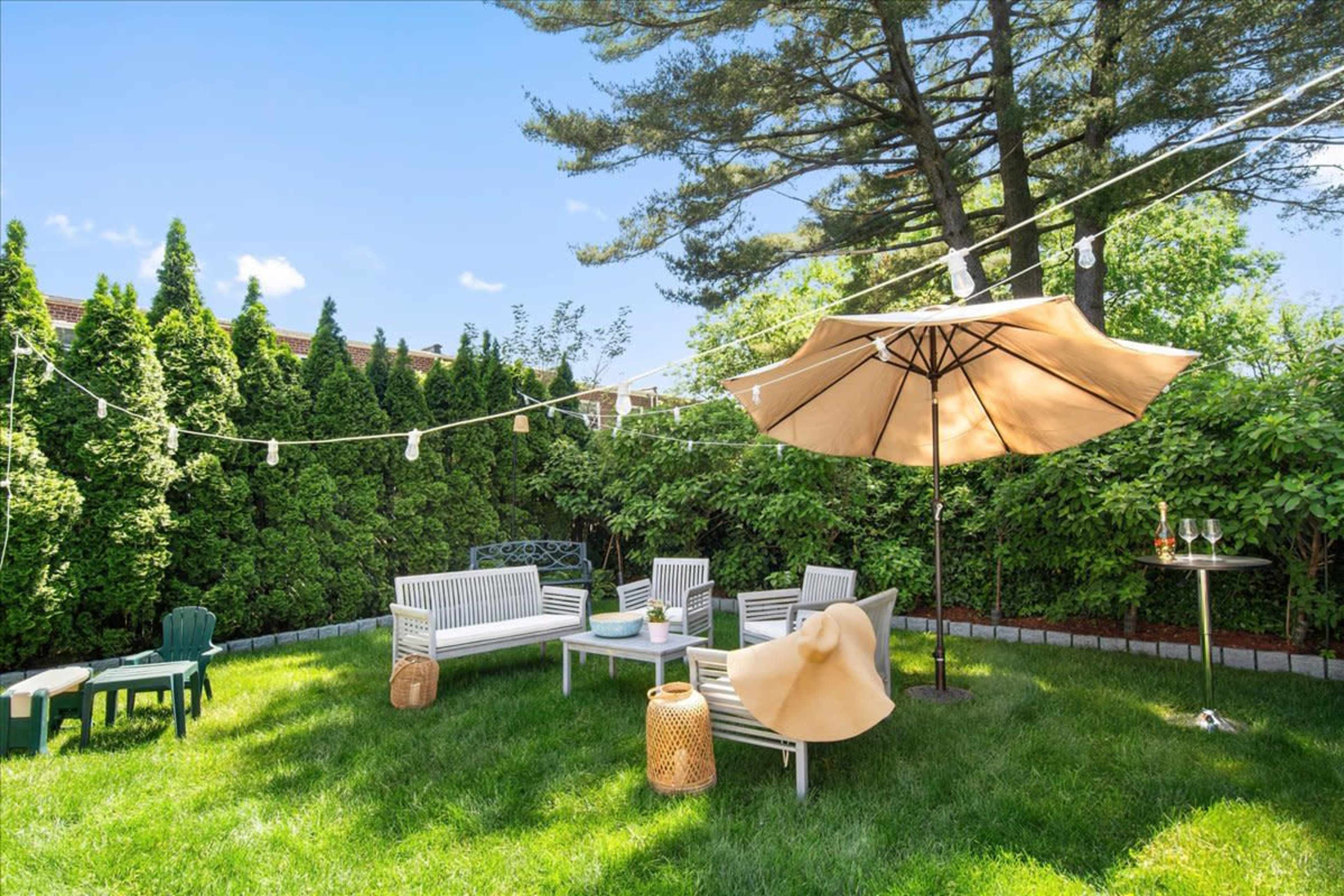 The image shows a neatly arranged backyard with seating, an umbrella, and string lights, surrounded by greenery.