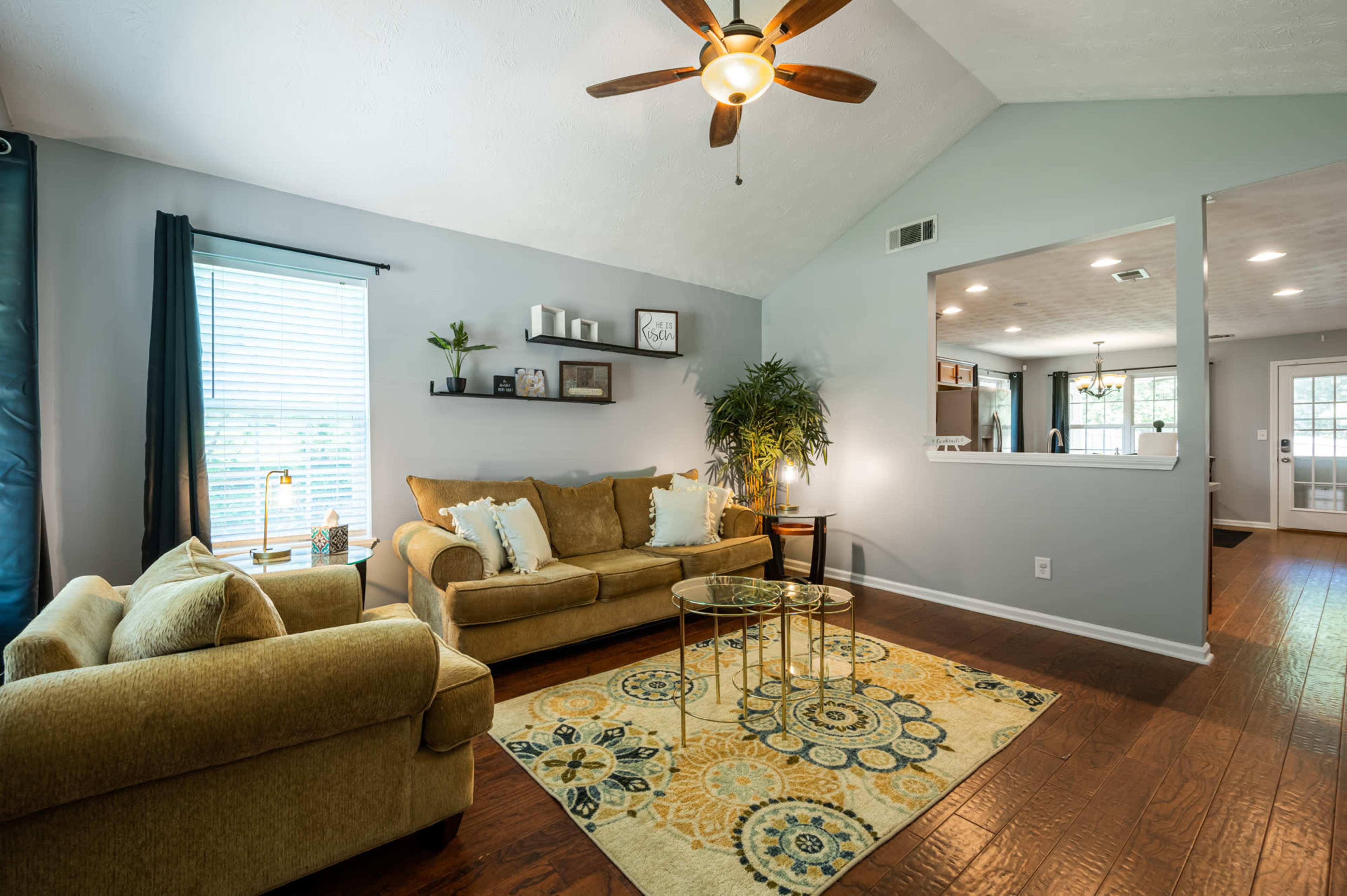 The living room features a tan sofa and loveseat, a decorative area rug, a ceiling fan, and a view into an adjoining kitchen.