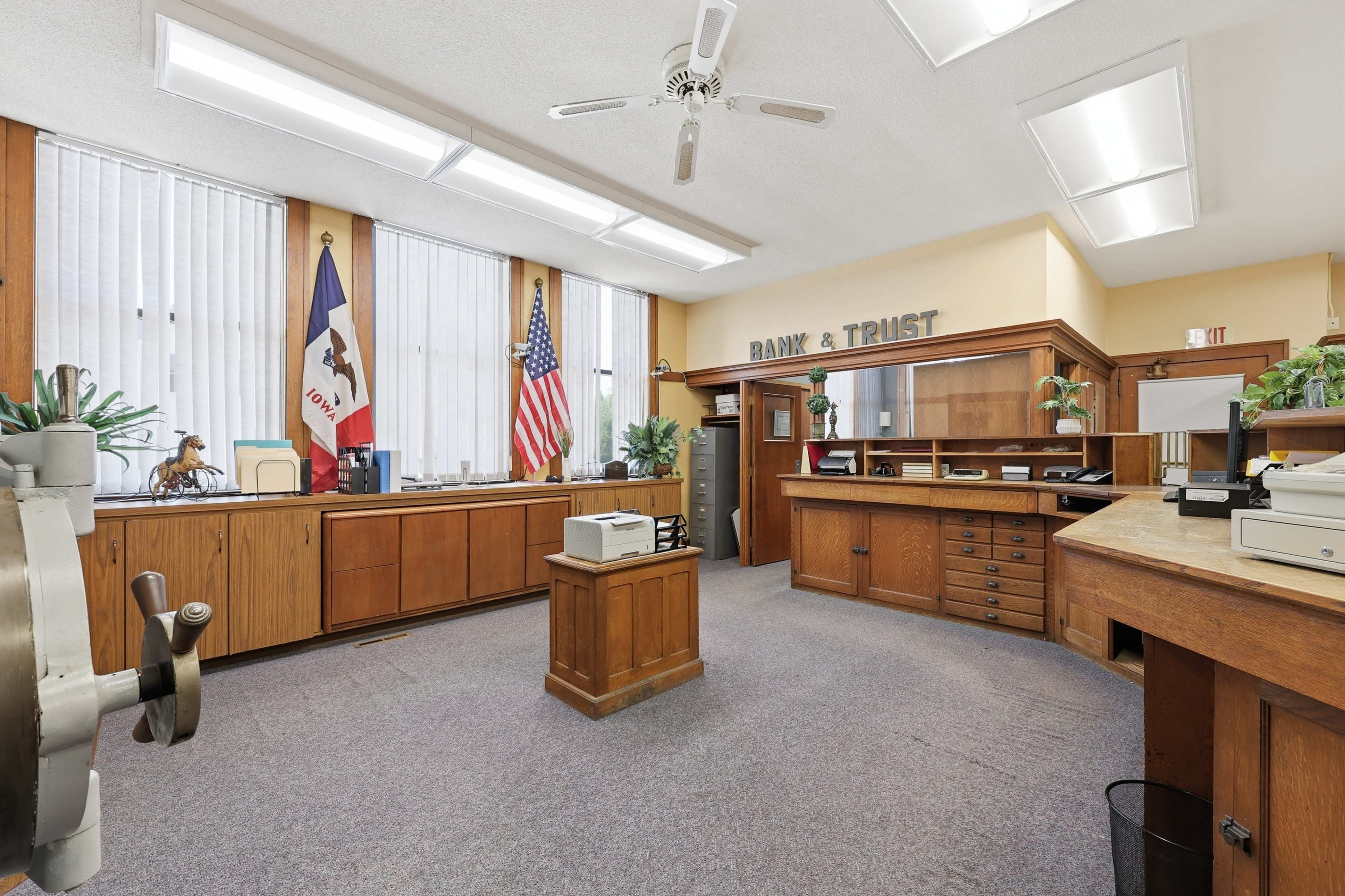 The image shows the interior of a bank office featuring wooden furniture, a vault door, and flags in a well-lit space.