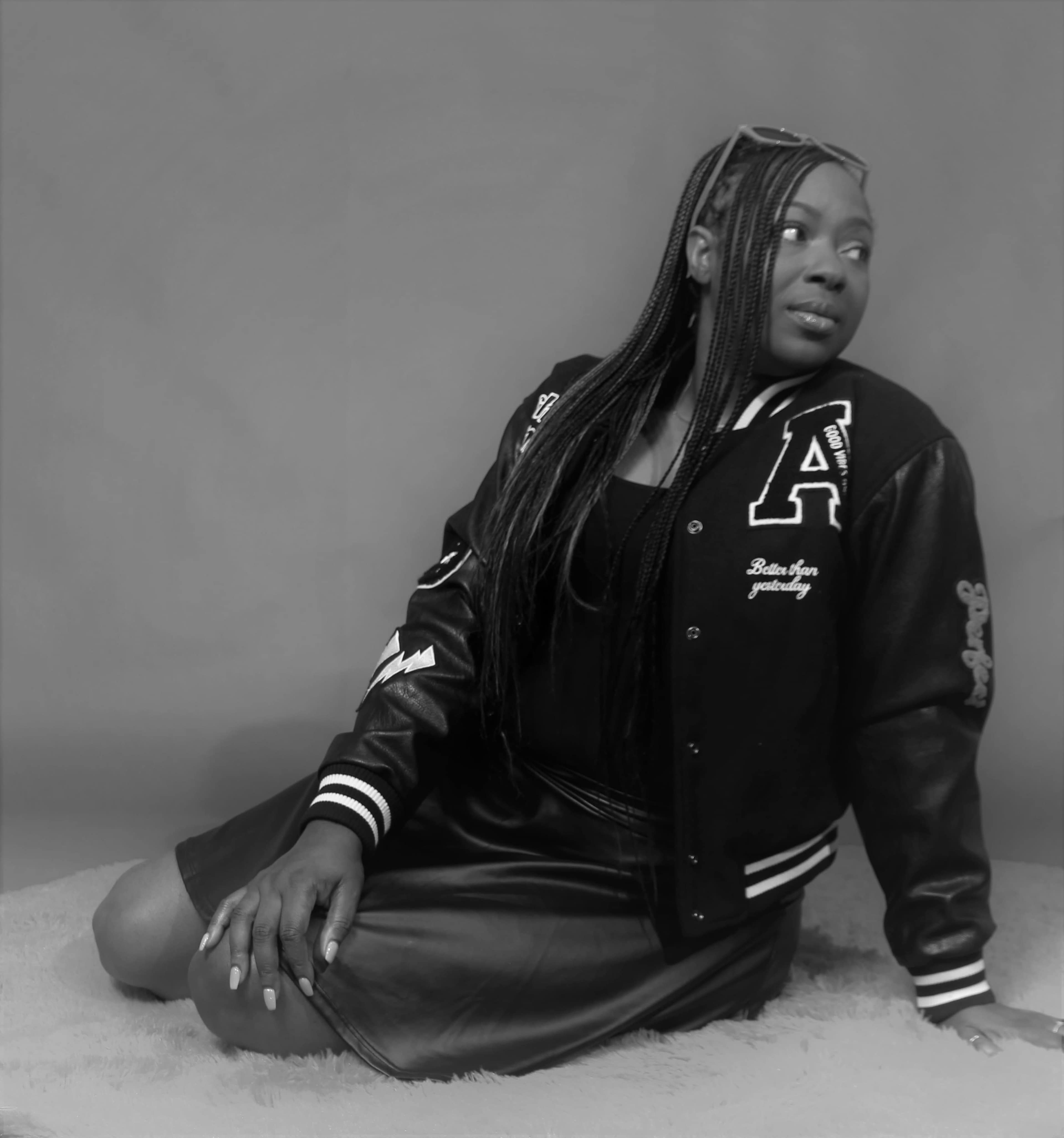 A person in a black leather jacket and skirt is sitting on a soft surface while looking over their shoulder.