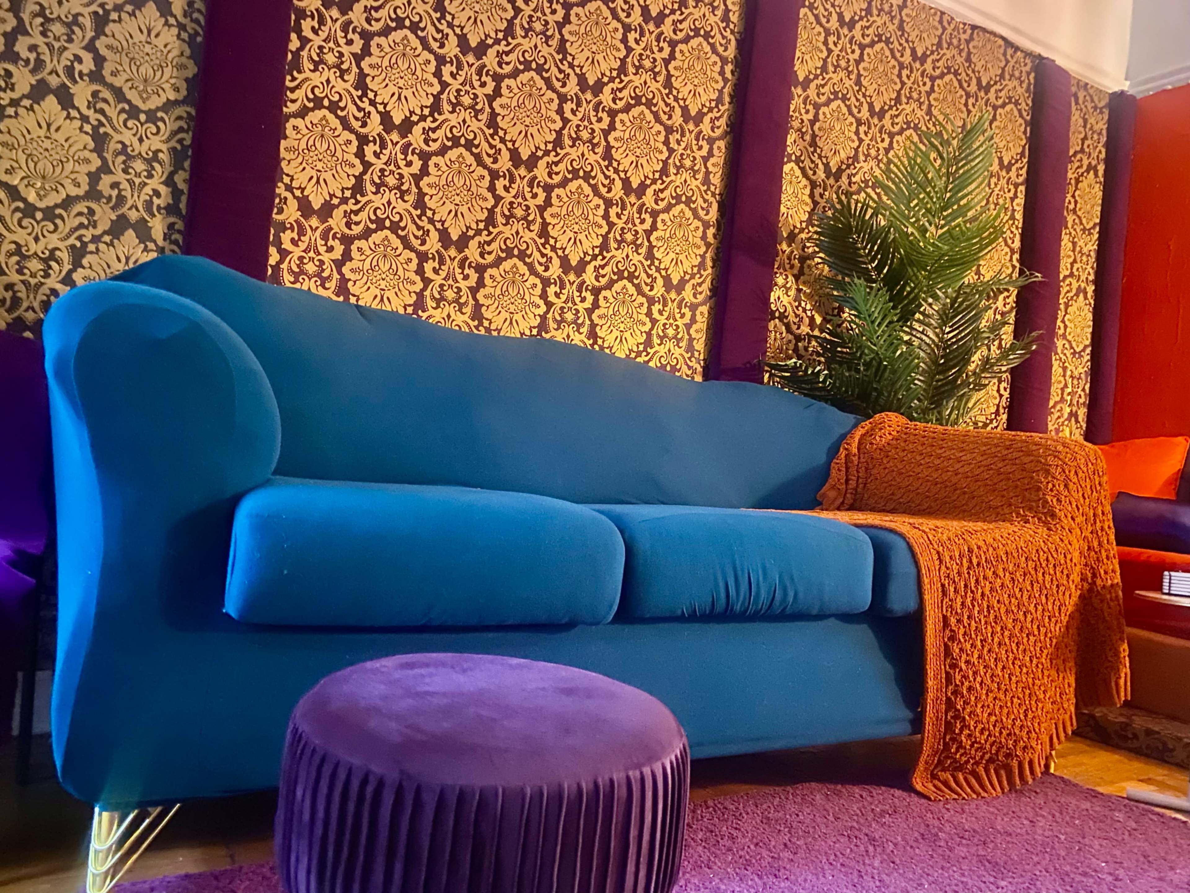 A turquoise sofa with an orange blanket rests against a patterned wall, accompanied by a purple ottoman and a potted plant.