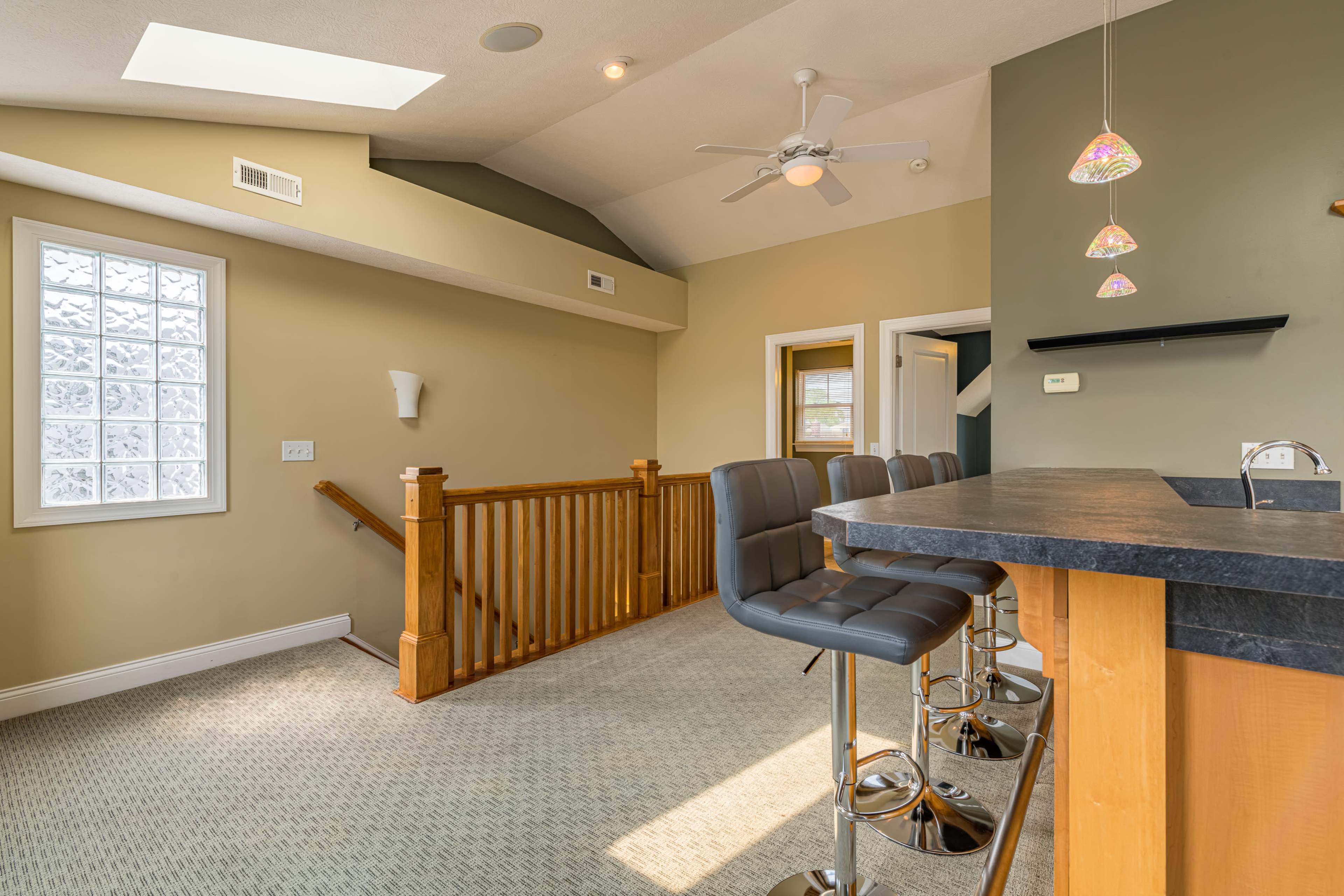 A well-lit room with a bar area featuring high stools, overlooking a staircase with wooden railings and a ceiling fan above.